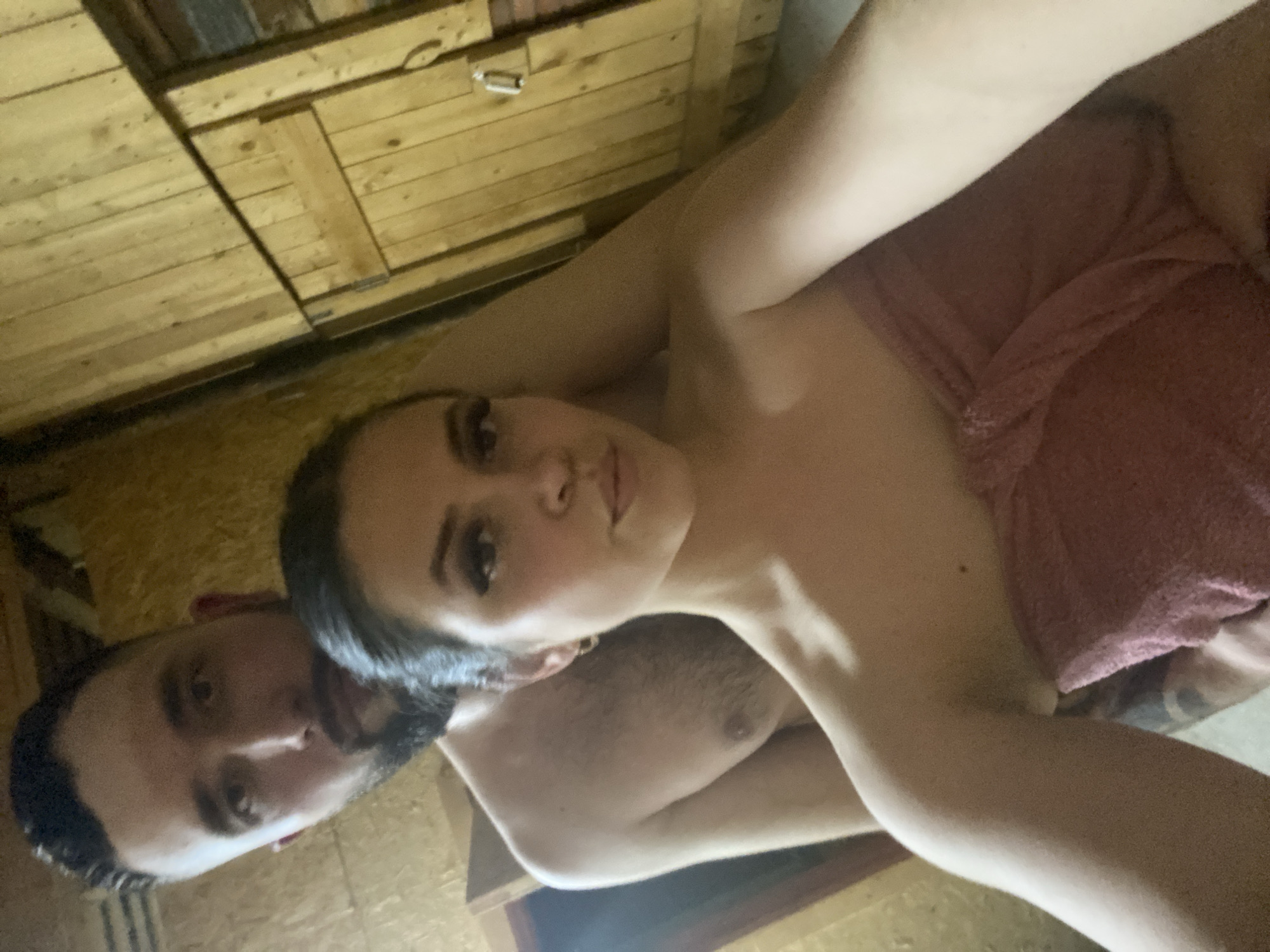 A selfie depicts a man and a woman in what appears to be a sauna. The woman is partially covered with a towel. The background is a wooden interior. The specific location in Zhigalki, Russia, is not identifiable from the image.
