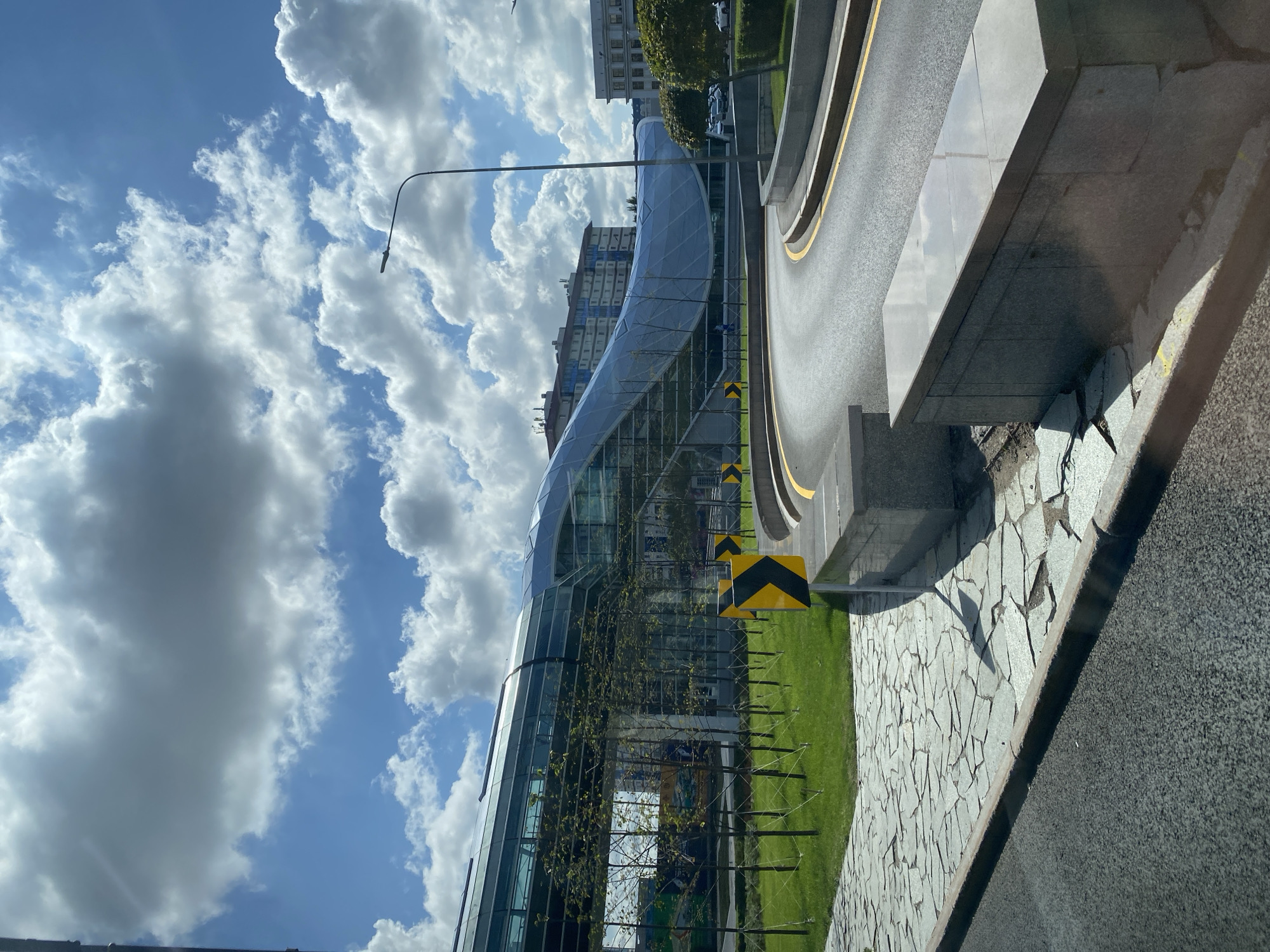 The image depicts a modern architectural structure with a curved glass facade, resembling a walkway or a building extension, situated in Astana, Kazakhstan. The building is set against a bright blue sky with scattered white clouds. In the foreground, a curved road with yellow lane markings is visible. On the left side of the road, a pathway made of irregular stone slabs leads to a grassy area. Several young trees with thin trunks are planted in the grassy area, supported by visible guy wires. Yellow and black directional signs indicating a sharp curve in the road are placed along the roadside. In the background, apartment buildings are visible. The overall scene suggests an urban environment with contemporary design elements and landscaping.