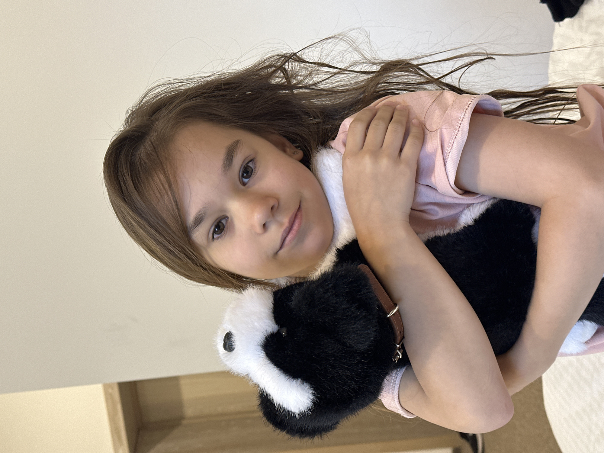 A young girl with long, dark brown hair is lying down and holding a stuffed animal that resembles a skunk. She is wearing a light pink shirt. The stuffed animal has black and white fur and appears to be a toy. The girl is looking towards the camera with a gentle smile. Her arms are wrapped around the stuffed animal. The background is a plain, light-colored wall, suggesting an indoor setting. The lighting is soft and natural. There is no visible text in the image. The overall mood is one of comfort and affection towards the toy.