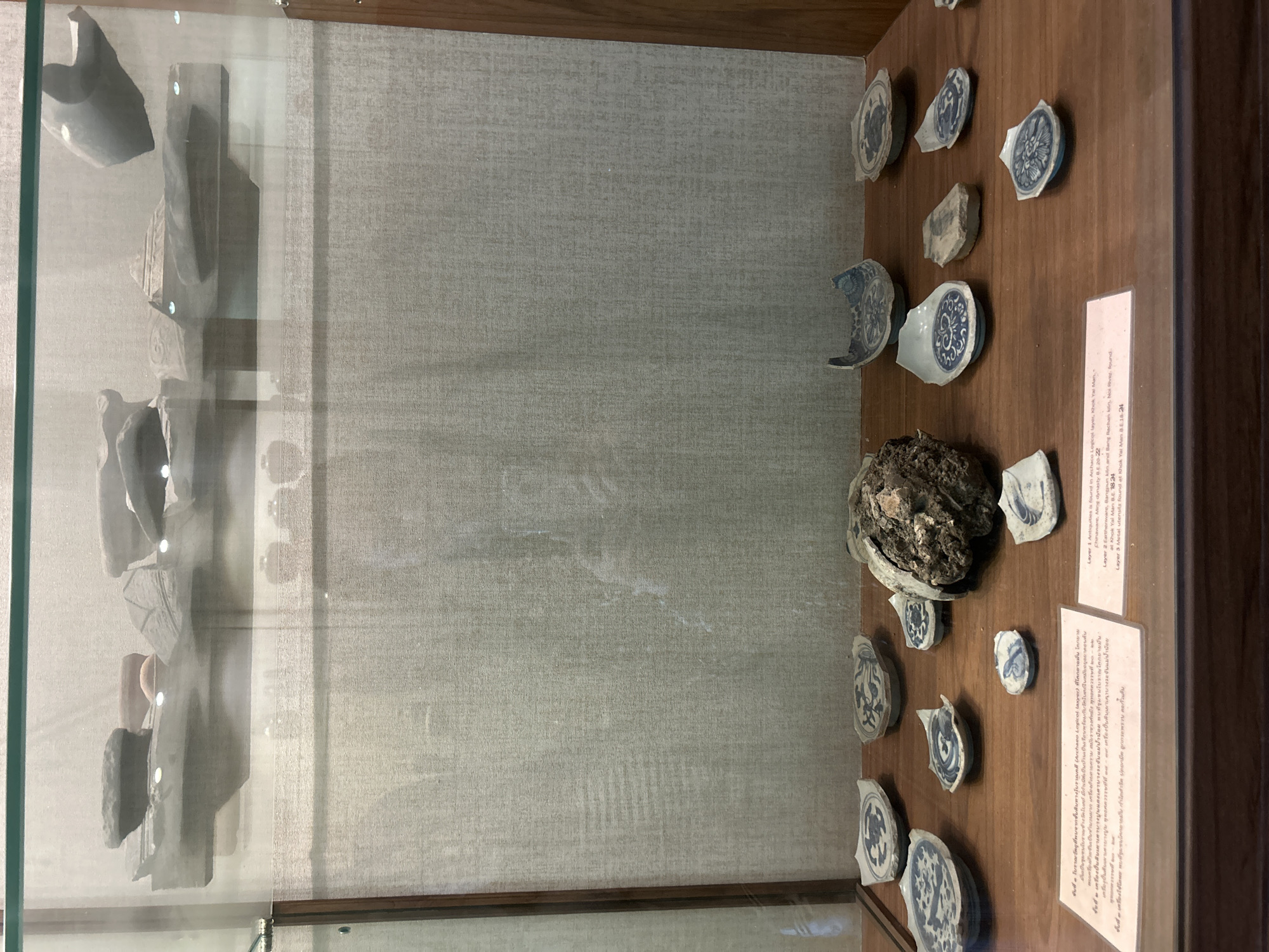 The image depicts an exhibition display case containing archaeological artifacts, likely within the city of Thai Ko Subdistrict, Thailand. The display features several layers of artifacts presented on wooden shelves. The top shelf holds fragments of pottery or stoneware, some appearing as large, rough pieces. The middle section of the display case is largely empty, with a textured fabric backdrop. The bottom shelf showcases numerous broken ceramic shards, primarily fragments of blue and white porcelain with intricate patterns. These shards are arranged alongside a substantial, irregular clump of earth or rock with embedded debris. A descriptive placard is positioned in front of the bottom shelf, providing information in both Thai and English about the provenance and dating of the artifacts, specifying their discovery in archaeological layers at Khok Yai Man and Bang Rachana, dating from B.E. 20-22 and B.E. 18-24 respectively. The display is housed within a glass-fronted cabinet with wooden framing. No people are visible in the image.