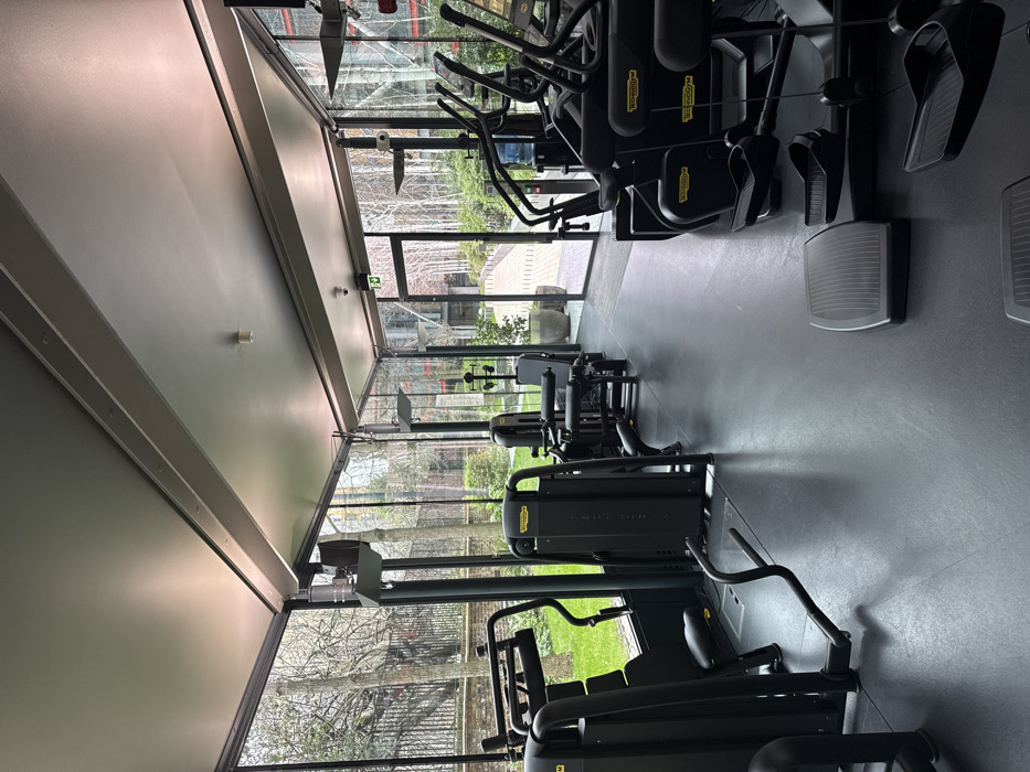 The image displays an unoccupied modern gym facility located within a residential or commercial development in London, United Kingdom. The room features a dark grey rubber floor and a light grey ceiling with exposed structural beams. The exterior walls are composed of floor-to-ceiling glass panels with dark grey framing, providing a view of the outdoor environment.
Within the gym, several pieces of black "Technogym" exercise equipment are visible. On the left side, resistance machines, including a chest press and a leg extension/curl machine, are positioned. Towards the right, a row of cardio machines, identifiable as ellipticals or treadmills by their foot pedals and upright handles, lines the glass wall. A blue water cooler is situated behind one of these cardio machines. A grey rectangular aerobic step is on the floor in the foreground, and a black, L-shaped metal bar lies flat on the floor in the mid-ground. An emergency exit sign (green with a white running man pictogram) is mounted above a glass door leading outside.
Through the glass, the exterior shows a landscaped area with numerous deciduous trees, mostly bare of leaves, suggesting autumn or winter. Green grass is visible on the left, alongside a low black metal fence. A paved walkway or bridge leads away from the building towards other modern structures in the distance, flanked by planters containing small trees or shrubs. No individuals are present in the scene.