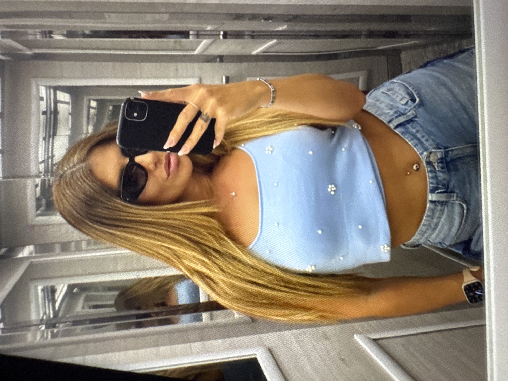 A young woman with long blonde hair is taking a mirror selfie in an elevator. She is wearing a light blue crop top adorned with small white beads and denim shorts. She has on sunglasses and is holding a black smartphone in her right hand. She is also wearing a silver bracelet on her left wrist and has a belly button piercing. The elevator walls appear to be mirrored and made of stainless steel. Based on the visual cues, the location is highly likely to be an elevator in a modern building, potentially a hotel or residential complex. There is no specific information within the image that allows for the precise determination of the city being Plovdiv, Bulgaria.
