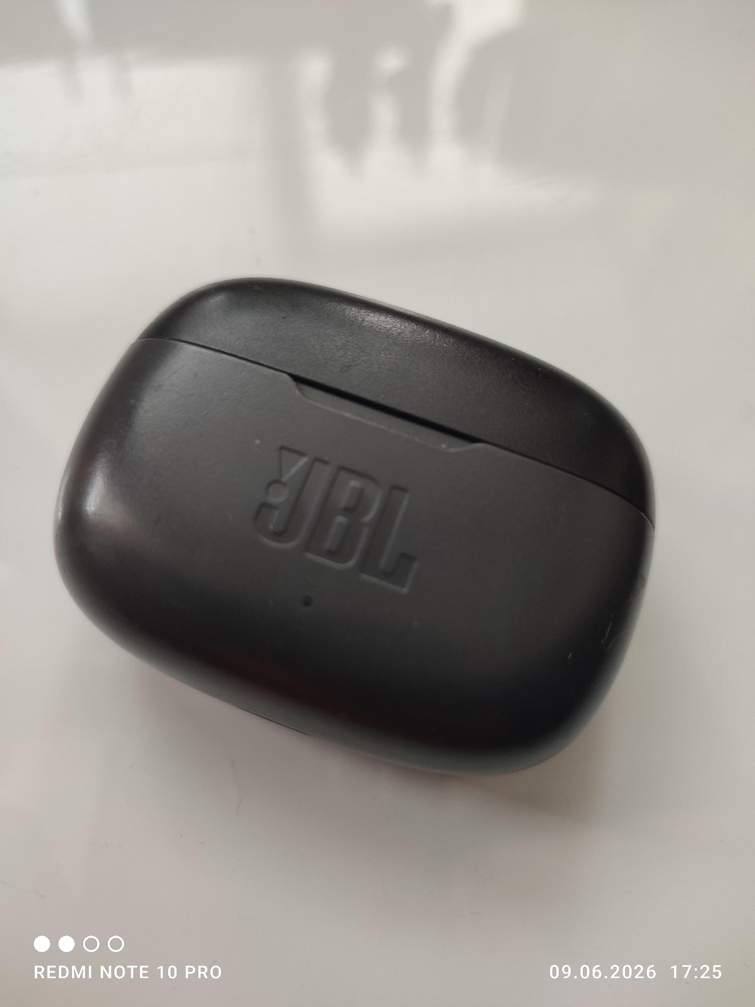 A black JBL charging case for wireless earbuds is shown from a high-angle, slightly overhead perspective. The case is closed and has the embossed JBL logo on its front. The surface of the case appears slightly textured and shows some minor scuff marks. The case rests on a white, possibly reflective surface, which is illuminated by soft, diffused light, casting subtle shadows. The background is out of focus and appears to be a bright, neutral color. The image is a close-up of the product.