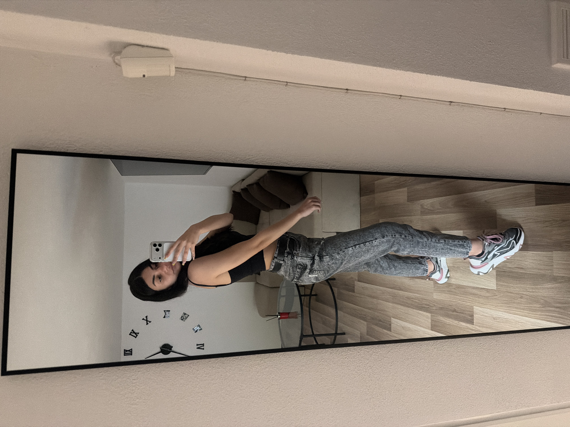 A full-length mirror selfie captures a young woman standing in an indoor room, identified as being in Gabrovo, Bulgaria. The woman has dark hair and is wearing a black tank top, acid-wash gray jeans, and athletic shoes with black, white, and pink accents. She is holding a white or silver smartphone with multiple camera lenses in her right hand, positioned to take the photo in the mirror. Her expression is neutral, focused on the camera.
The room reflected in the mirror features light-colored walls and a light wood-look laminate floor. Behind the woman, visible in the reflection, is a sofa with dark brown cushions and a small round glass-top table with a black metal frame. On the wall behind the woman, a decorative wall clock with black hands and individual Roman numeral markers (III, IV, IX, X, XI, XII are clearly visible) is mounted.
Above the mirror, a white rectangular device, possibly a smoke detector or a sensor, is attached to the ceiling. A thin wire or cable also runs along the ceiling/wall line above the mirror. The lighting suggests an indoor setting, likely not illuminated by strong natural daylight.