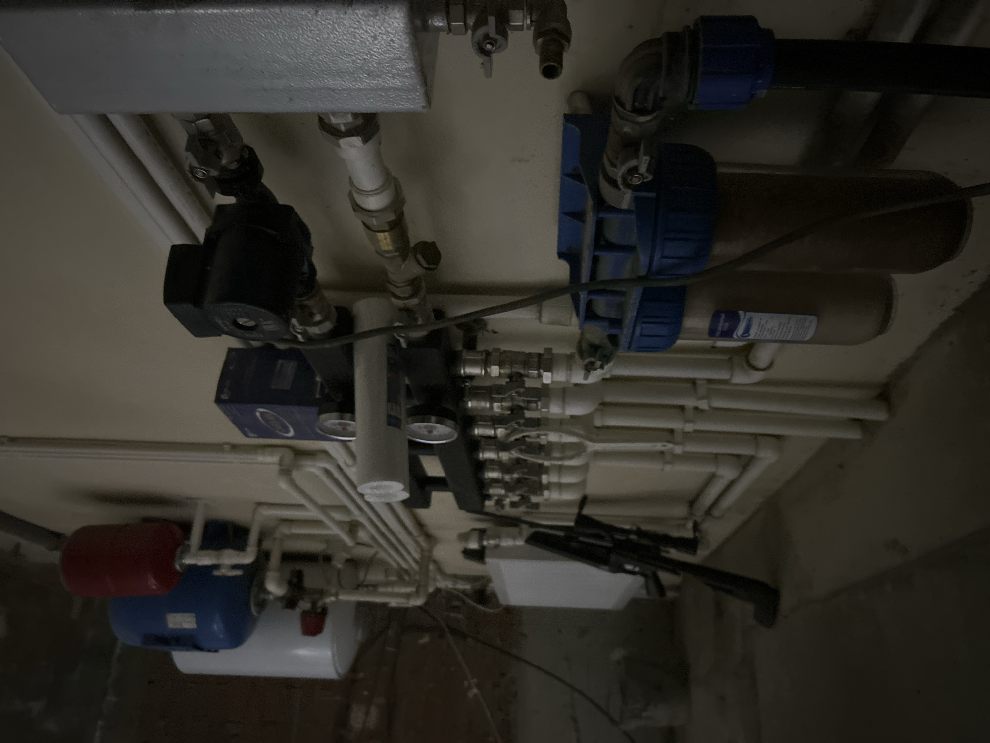 The image shows a utility area, possibly a basement or mechanical room. Various plumbing components are visible, including pipes, valves, filters, a pressure tank (red), a water heater (white), and a water softener (blue). A rifle is mounted on the wall. The image lacks information about people, subjects, actions, interactions, or the specific location within Noviy, Russia.