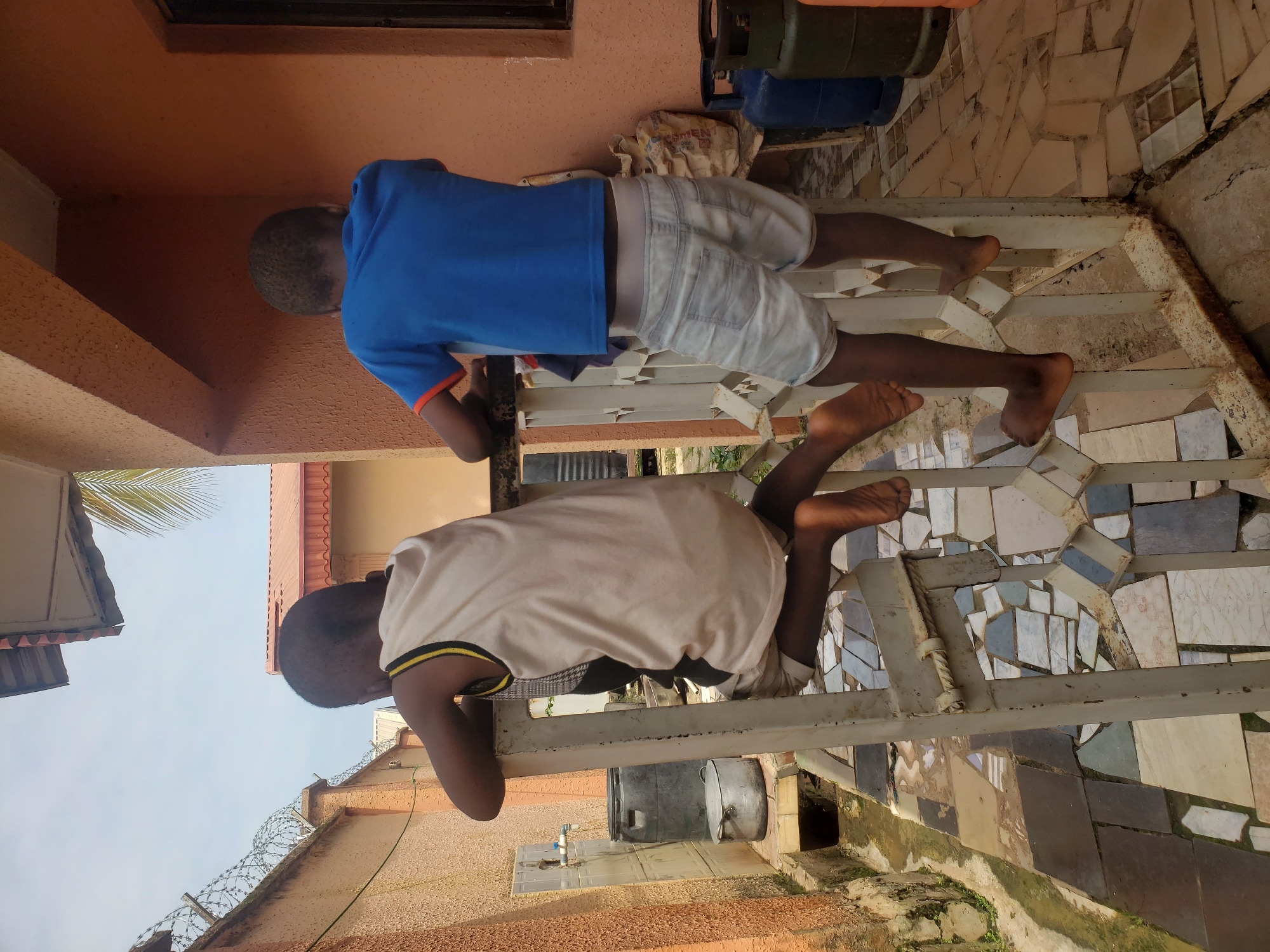 In Sabon Tasha, Nigeria, two young boys are seen from behind, standing on a white metal railing. The boy on the left is wearing a light-colored sleeveless shirt with black and yellow trim and light-colored shorts. He is perched on the railing, facing away from the camera. The boy on the right is wearing a blue t-shirt with orange trim and light-colored denim shorts. He is also standing on the railing. The environment appears to be an outdoor space adjacent to a building with an orange-colored wall. In the background, there are gas cylinders, a large plastic barrel, a metal pot, a faucet on a tiled wall, and a narrow drain. The ground is covered with a mosaic of irregularly shaped tiles. The sky is visible above, with a palm frond from a tree extending into the frame. A barbed wire fence runs along a wall in the distance.