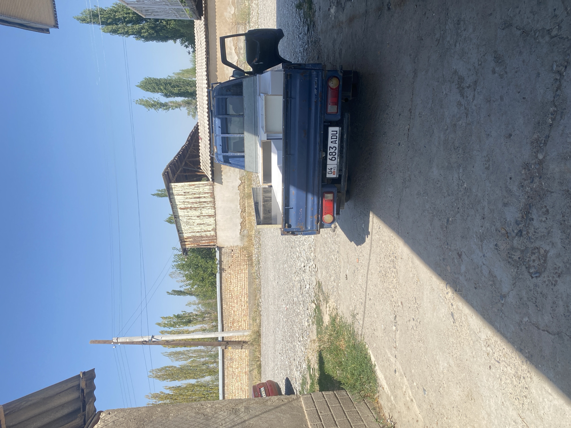 A blue pickup truck is parked on a dirt and gravel surface in an outdoor area with a brick wall and some trees in the background. The truck's bed is loaded with several white rectangular objects, possibly boxes or furniture pieces. The driver's side door of the truck is open, and a license plate with the characters "04 KG 683 ADU" is visible on the rear of the vehicle. The area appears to be a residential or industrial space, with a corrugated metal roof structure and a utility pole also visible. The sky is clear and blue.