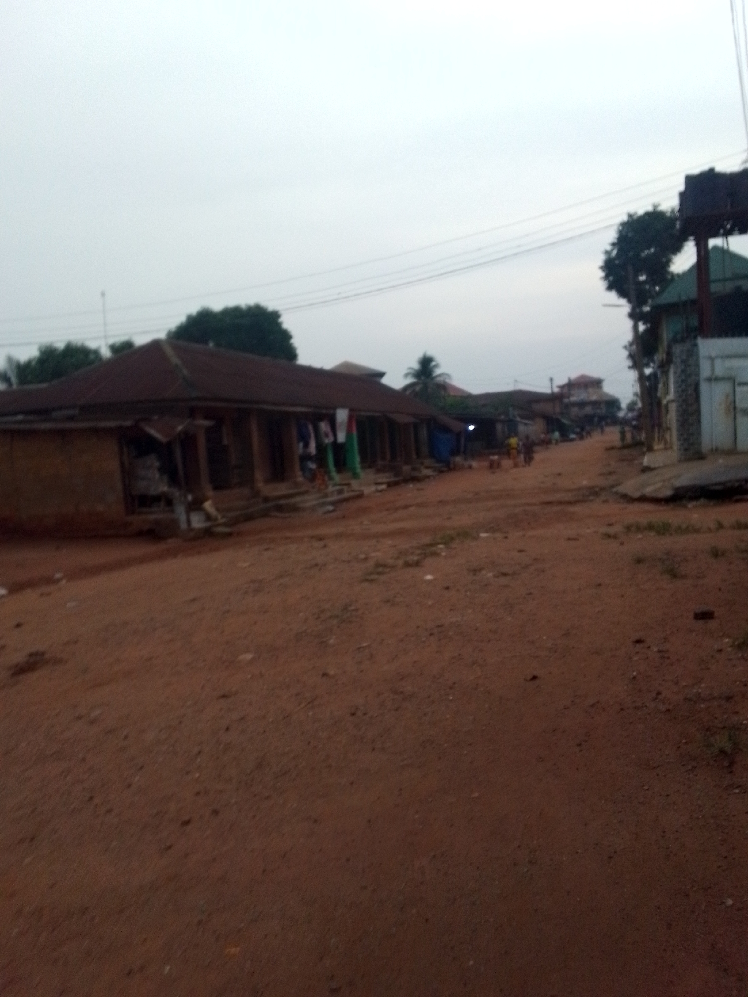 This image captures a street scene in what appears to be a rural or semi-urban area, consistent with Orlu, Nigeria. The dominant feature is a wide, unpaved road made of reddish-brown dirt or laterite, stretching into the distance. The road surface shows variations in texture and some small patches of sparse greenery.
On the left side of the road, a long, low-slung building with a dark, corrugated metal roof and what appears to be unpainted earth or cement walls stands prominently. It features multiple open doorways or shopfronts along its facade. Colorful fabrics or clothing are seen hanging outside one of these openings, suggesting a market or retail activity. Further down the street on the left, other buildings and structures, some partially obscured, continue to line the road.
On the right side of the road, closer to the foreground, a building with contrasting lighter-colored walls and a textured, possibly stone-clad section is visible. Above it, a dark, elevated structure, potentially a water tank stand or a billboard frame, is silhouetted against the sky. Power lines or utility wires traverse the upper part of the frame.
The sky is uniformly overcast and light grey, casting a dim, diffused light over the entire scene. This suggests either early morning, late afternoon, or a heavily clouded day. A few indistinct figures of people can be seen further down the road, walking or standing, indicating some daily activity. Trees and foliage are scattered throughout the background, adding to the natural setting. There is no discernible text visible in the image.