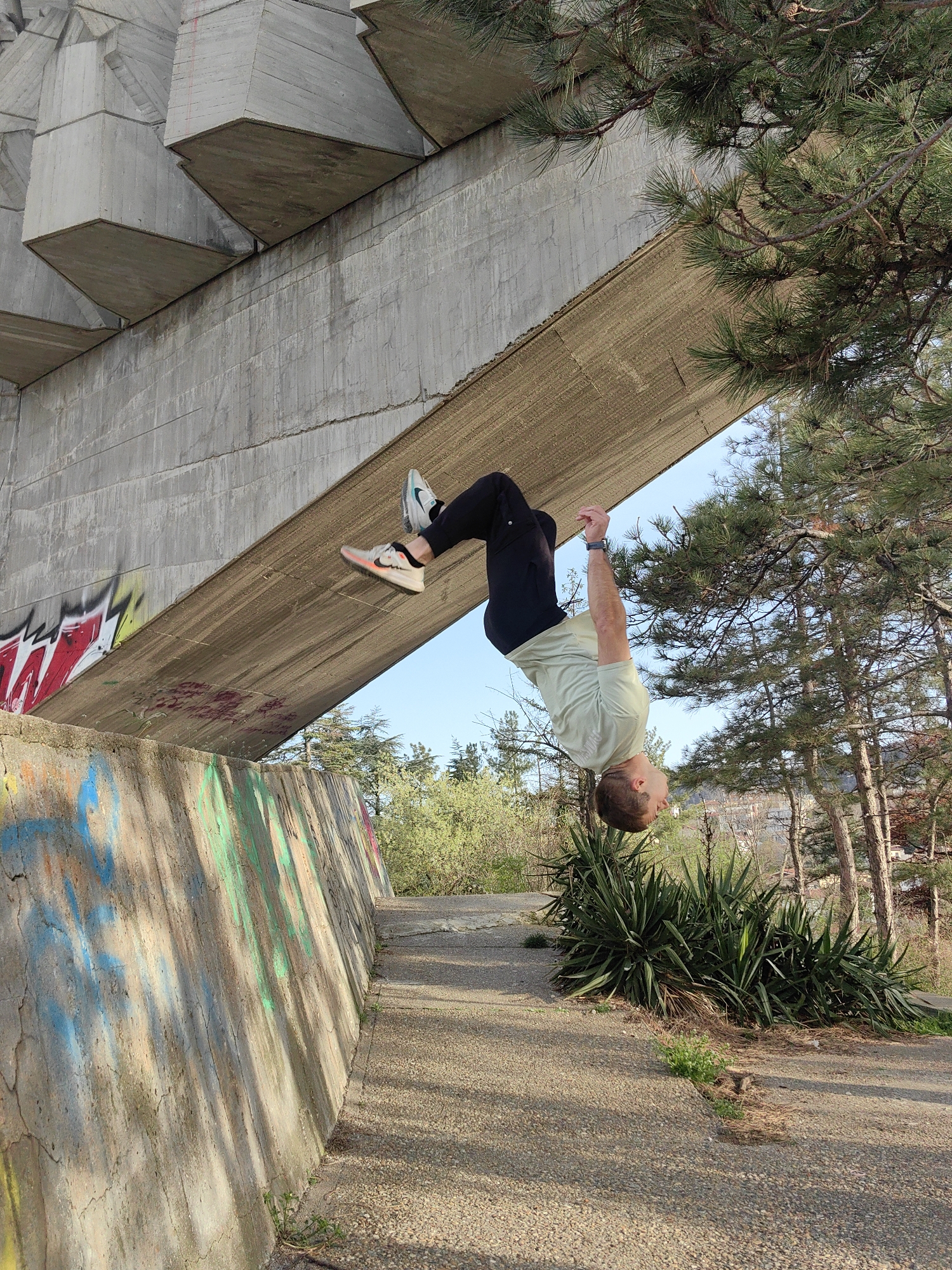 A man in black pants, a pale green t-shirt, and athletic shoes is hanging upside down under a concrete structure. The location appears to be an urban park setting in Varna, Bulgaria near the Monument of the Bulgarian-Soviet Friendship, evidenced by the visible concrete architecture. Graffiti is present on the walls. Trees and shrubs are visible behind the man. The ground is paved with small stones.