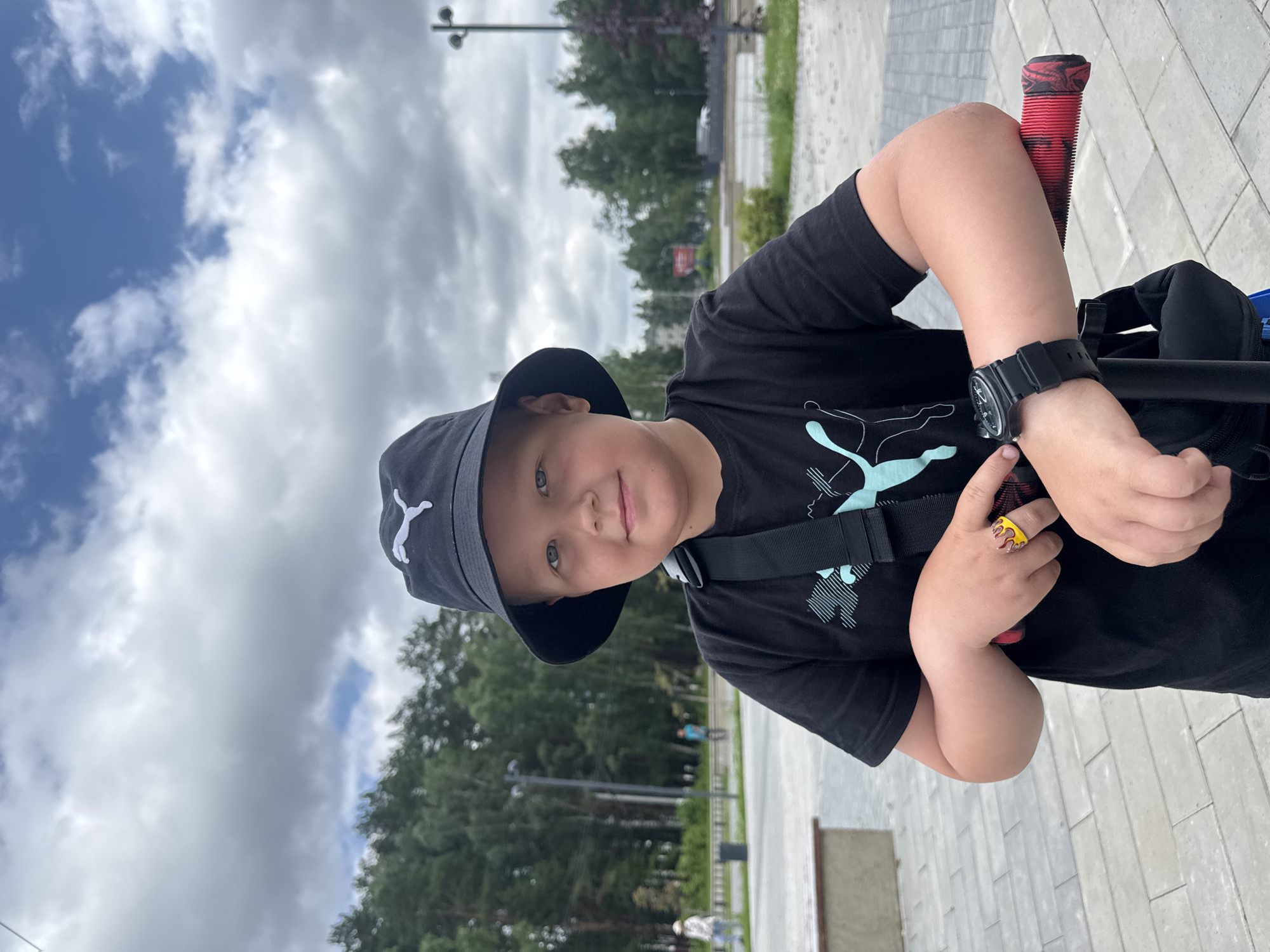 A young boy, wearing a black bucket hat with a white Puma logo and a black t-shirt with a blue graphic, poses for a photo outdoors. He has fair skin and light blue eyes, and is looking directly at the camera with a slight smile. The boy is holding onto the handlebars of a scooter, which have red grips. On his left hand, he wears a yellow ring with a red flame design, and on his left wrist, he wears a black wristwatch. The background shows a paved area with trees and a cloudy sky. The scene appears to be in an urban park or public space. The location is identified as the city of Chaikovsky, Russia.