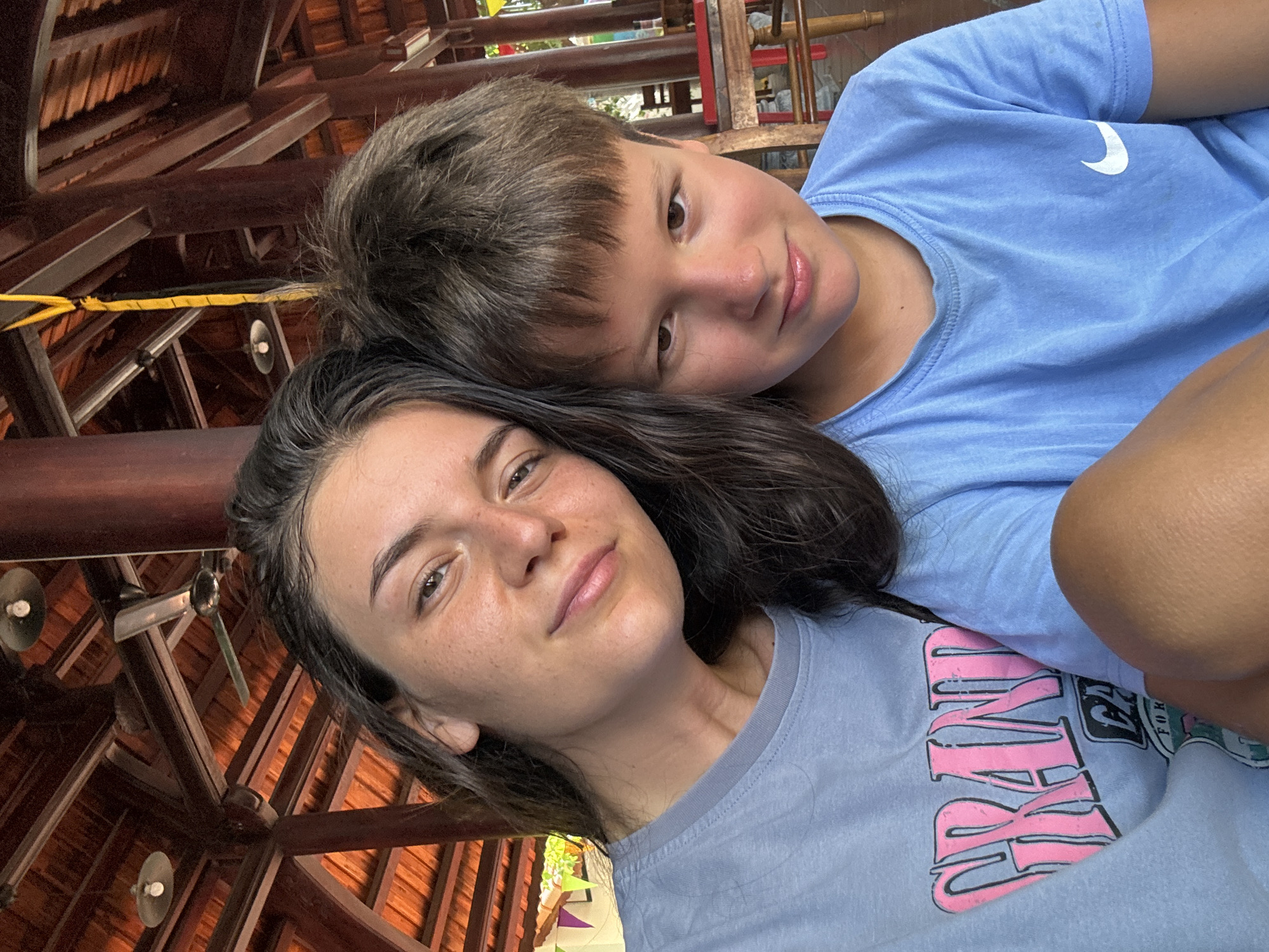 The image shows two people posing for a photo in what appears to be an indoor or covered outdoor space. The person in the foreground is a young woman with dark hair and a light complexion. She is wearing a gray t-shirt with pink lettering. The person behind her is a younger boy with short brown hair, wearing a light blue t-shirt with a white swoosh logo. The background shows a complex wooden structure with beams and decorative elements. The location is Mui Ne, Vietnam.