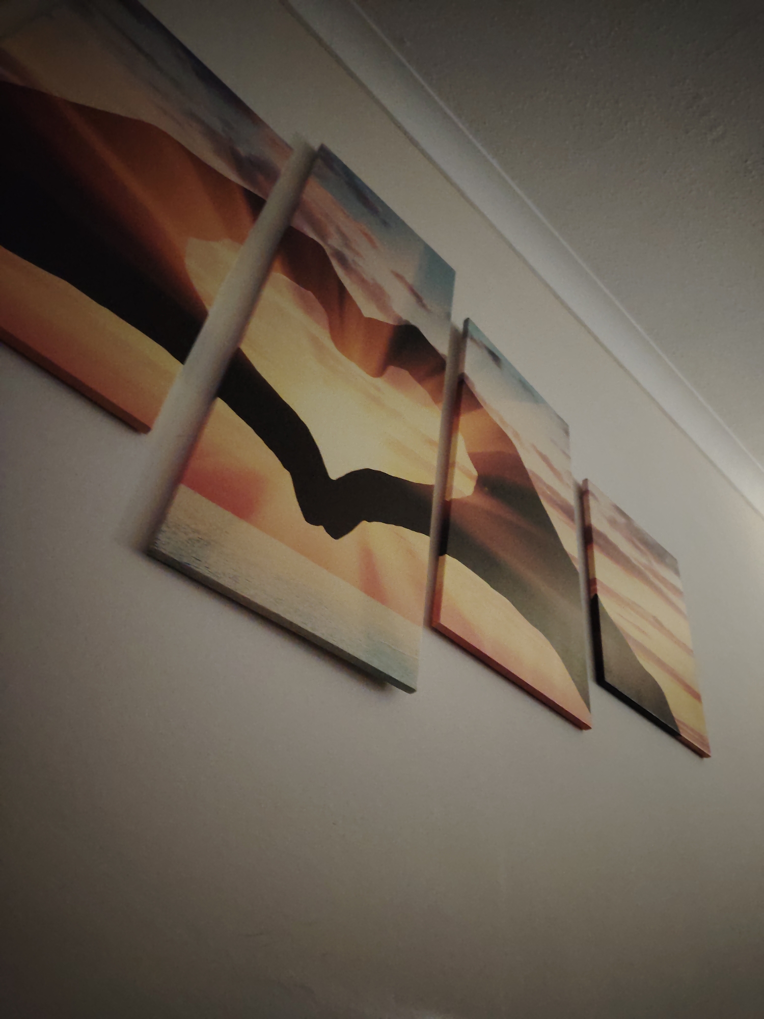 The image captures a set of five multi-panel canvas prints mounted on a light-colored wall in an indoor setting. The photograph is taken from a low, upward angle, showcasing the wall art and a section of the ceiling.
The artwork depicts a warm desert landscape, likely at sunrise or sunset, featuring undulating sand dunes in vibrant shades of orange, amber, and reddish-brown, contrasting with a cooler, light blue and grey sky. A prominent element within the central panels is the silhouette of two hands clasped together, creating a sense of connection or journey across the desert scene.
The five panels are arranged horizontally, with varying dimensions; the leftmost panel is the largest, and the panels progressively decrease in size towards the right, creating a dynamic, staggered display. The canvases appear stretched over frames, with glimpses of the edges visible.
Above the wall art, a white ceiling with a subtle coving or trim runs horizontally. The wall itself is painted a neutral, possibly light grey or off-white, color. The lighting in the room appears soft and ambient, allowing the warm colors of the artwork to stand out. There is no visible text in the image. The scene suggests a tranquil and contemplative atmosphere within a home in Huntingdonshire, United Kingdom.