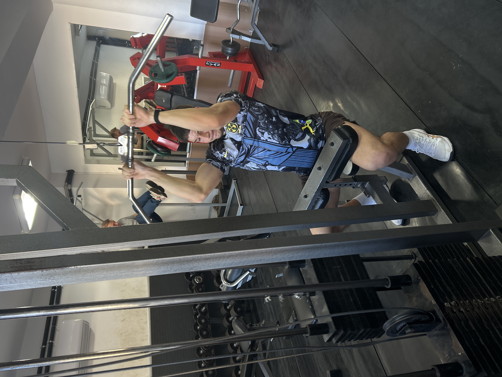 A man is captured in a gym setting, engaged in an exercise using a cable machine. The image is oriented sideways, showing the man lying on his back with his head towards the left, pulling a metal bar towards his chest. He appears focused, with a determined expression. He wears a dark and blue patterned short-sleeve t-shirt with a distinct logo, dark shorts, white athletic shoes with brown/gold accents, white socks, and a dark wristband on his left arm.
The gym features dark rubber flooring and light-colored walls. In the background, various pieces of gym equipment are visible. A large mirror reflects parts of the gym, including red-framed machinery with the text "LYXN" visible, an air conditioning unit, and another person lying on a bench. To the right of the main subject, a weight stack shows faintly visible numbers like 2, 4, 6, 8, 10, indicating the resistance level of the machine. A rack of dumbbells and other cable pulleys are also seen in the background. The overall scene depicts an active indoor workout environment in Zlatograd, Bulgaria, illuminated by artificial lighting.