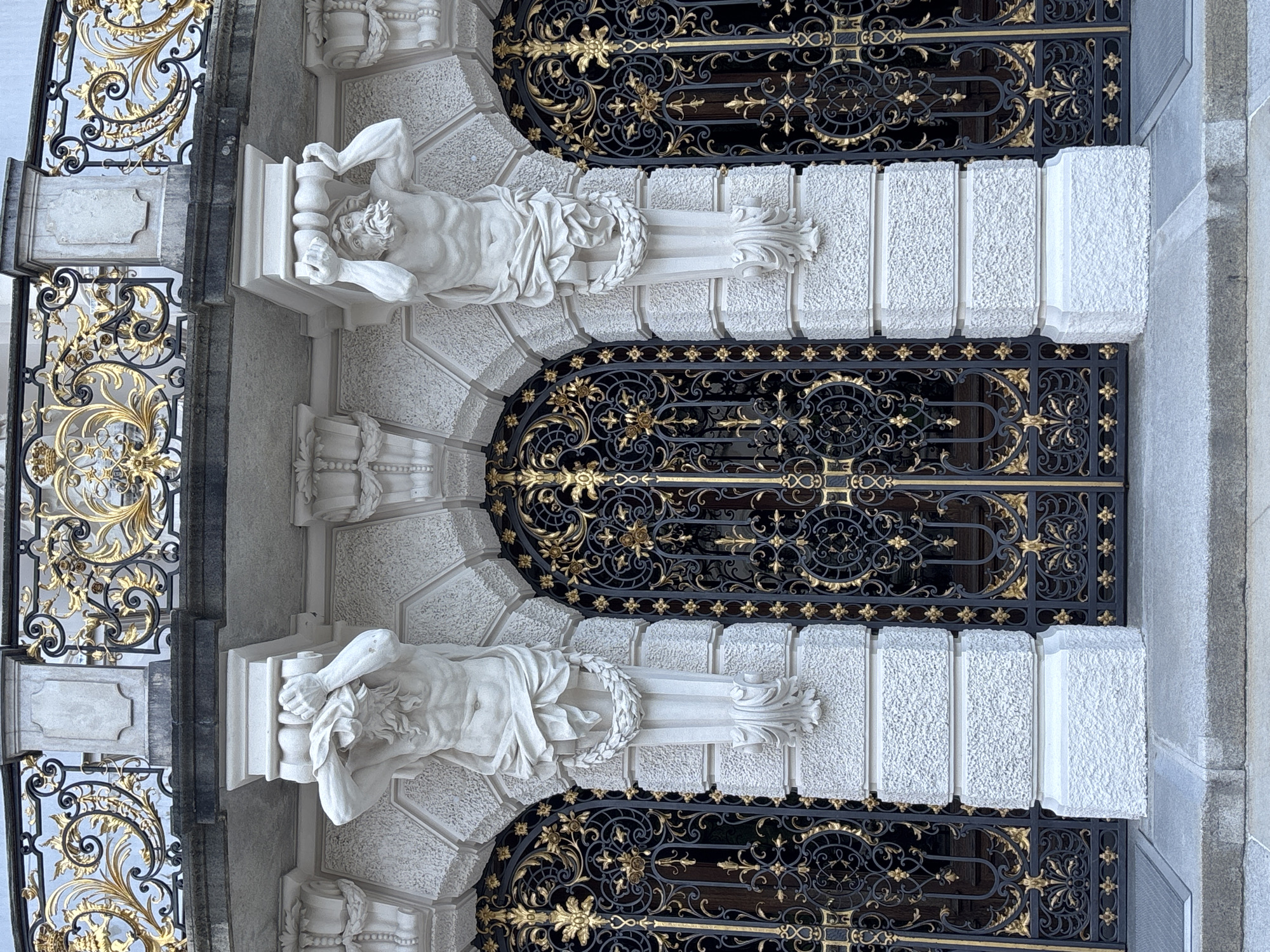 The image shows the exterior of a building, likely part of Ettal Abbey in Ettal, Germany. The architecture features ornate white stone sculptures of muscular male figures supporting ornate golden and black metal gates. These gates appear to be doors or entrances to the building, featuring intricate scrollwork and floral patterns. Above the sculptures and gates, a balcony or upper level is visible, also adorned with golden metal railings and decorative elements. The overall impression is one of grandeur and historical significance, with a focus on classical architectural and sculptural elements. The lighting suggests it is daytime, possibly overcast, casting soft shadows on the textured stone surfaces. There are no people visible in the image.
