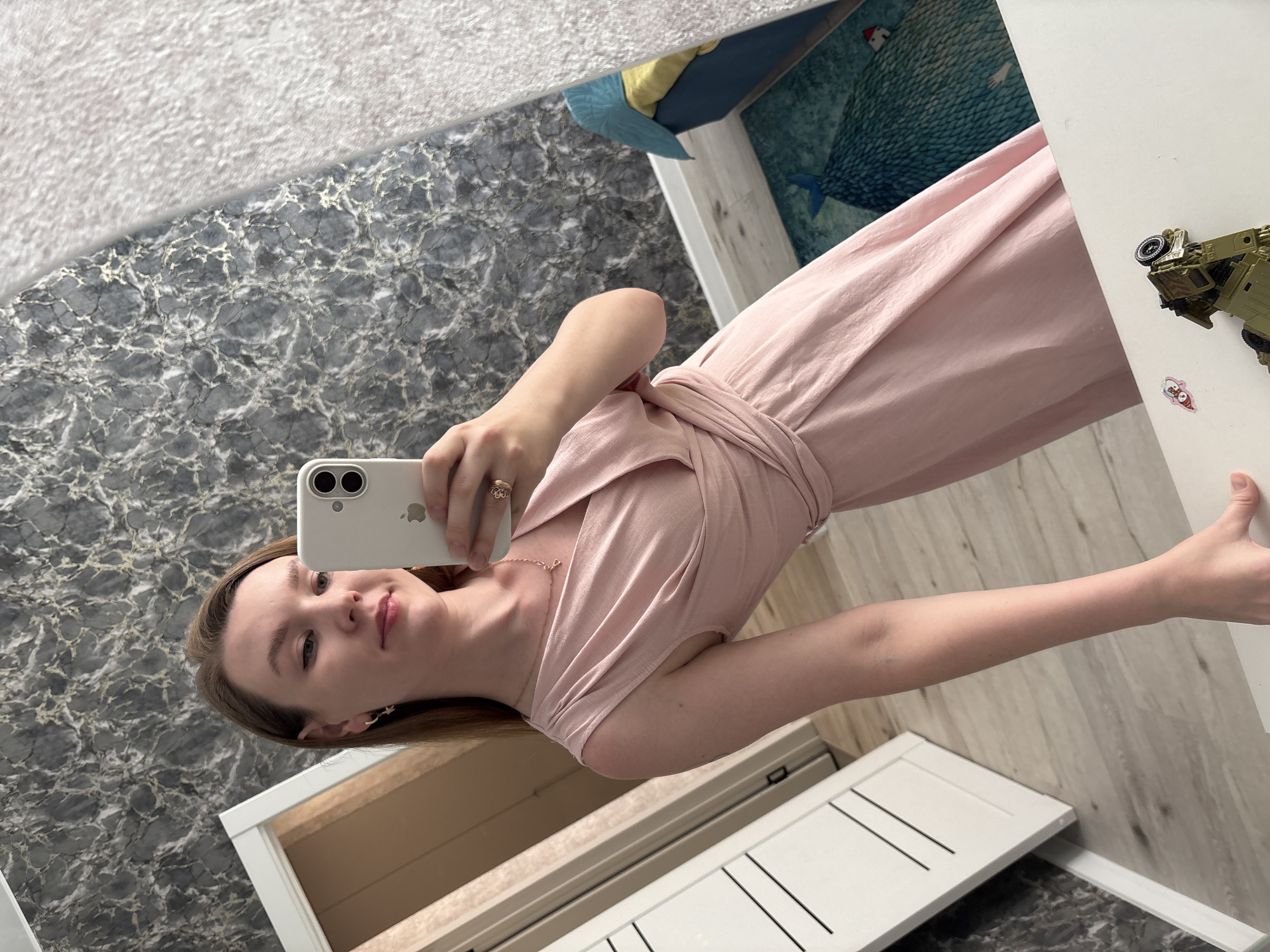 A young woman is taking a selfie in a mirror. She is wearing a light pink dress. The background shows a room with wallpaper, a piece of furniture, and a child's drawing or painting on the wall. A toy vehicle is on the furniture. The location is Izhevsk, Russia.