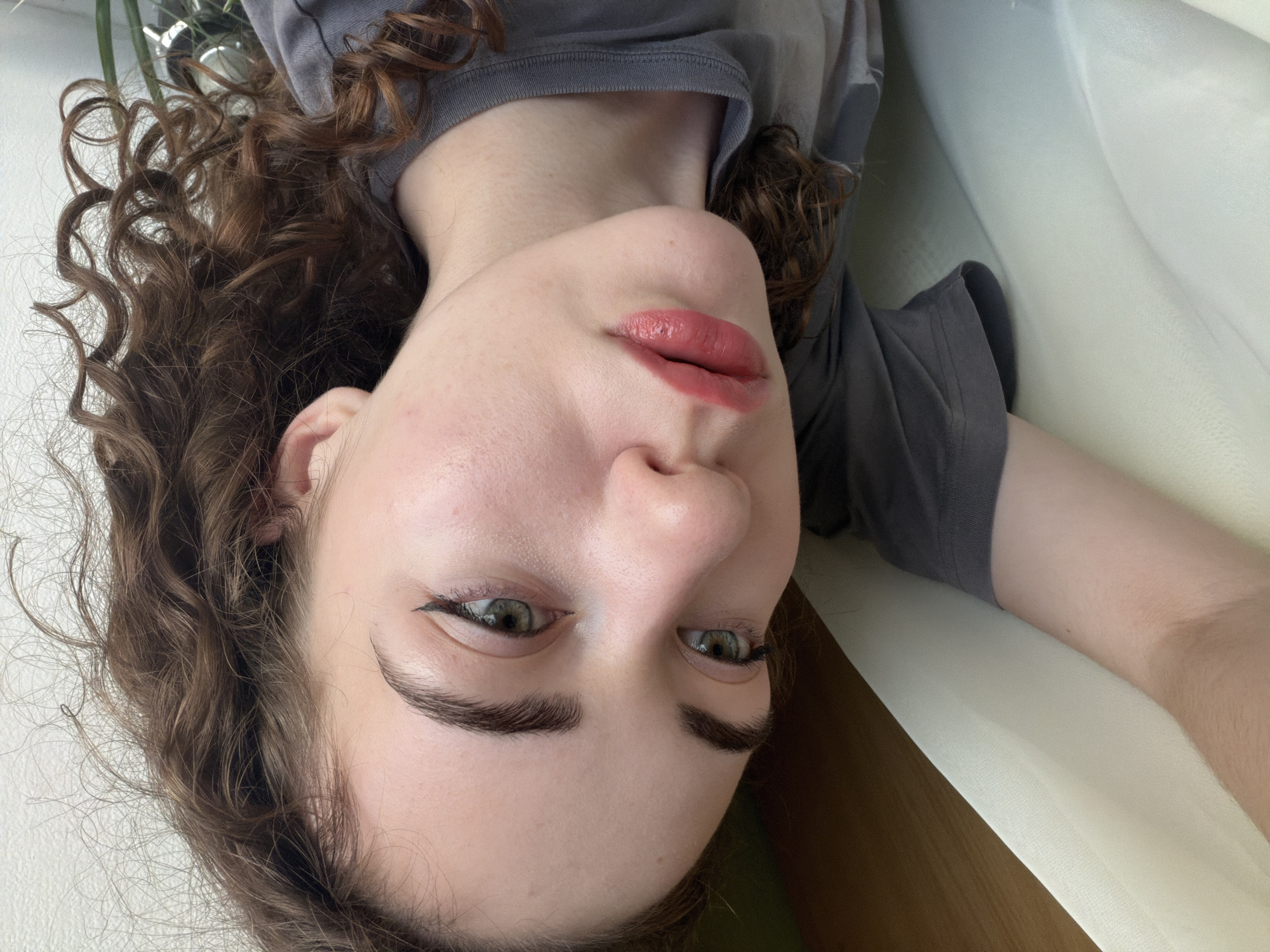 A close-up, rotated photograph shows a person's face and upper body. The person has curly, dark brown hair framing their face. Their eyes are green and their eyebrows are dark and well-defined. They are wearing makeup, including eyeliner and lipstick in a muted pink shade. The skin appears pale and smooth, with a hint of blush on the cheeks. The person is wearing a grey t-shirt. The background is out of focus, but appears to be a light-colored surface, possibly a sofa or bed, with some shadowing. The overall lighting suggests an indoor setting. There is no visible text in the image. The image was likely taken during the day, as indicated by the natural-looking lighting. The scene appears to be casual and personal, possibly a selfie. No specific activity or event is discernable, beyond the act of being photographed. The emotion conveyed is neutral to slightly thoughtful.