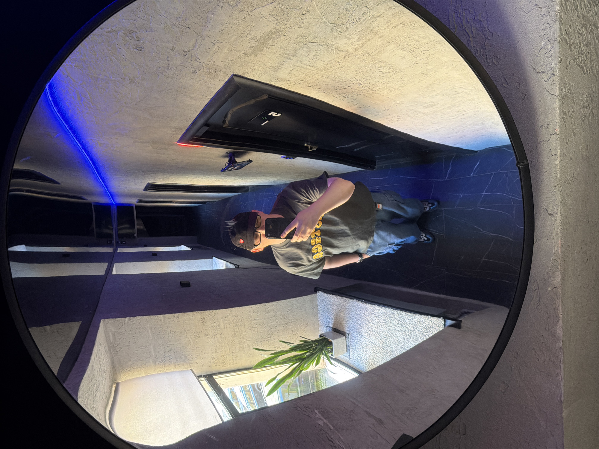 A person is taking a selfie in a fisheye mirror, reflecting a hallway. The person is wearing a dark t-shirt with yellow lettering, blue jeans, a black baseball cap with a red heart emblem, and glasses. They are holding a smartphone in their right hand. The hallway has textured walls, a window with blinds on the left, and several dark doors. Blue LED lights are installed along the top of the wall. The floor is tiled with a dark pattern. The scene is located in a commercial building, possibly a hotel or office, in the city of Saint Petersburg, Russia.