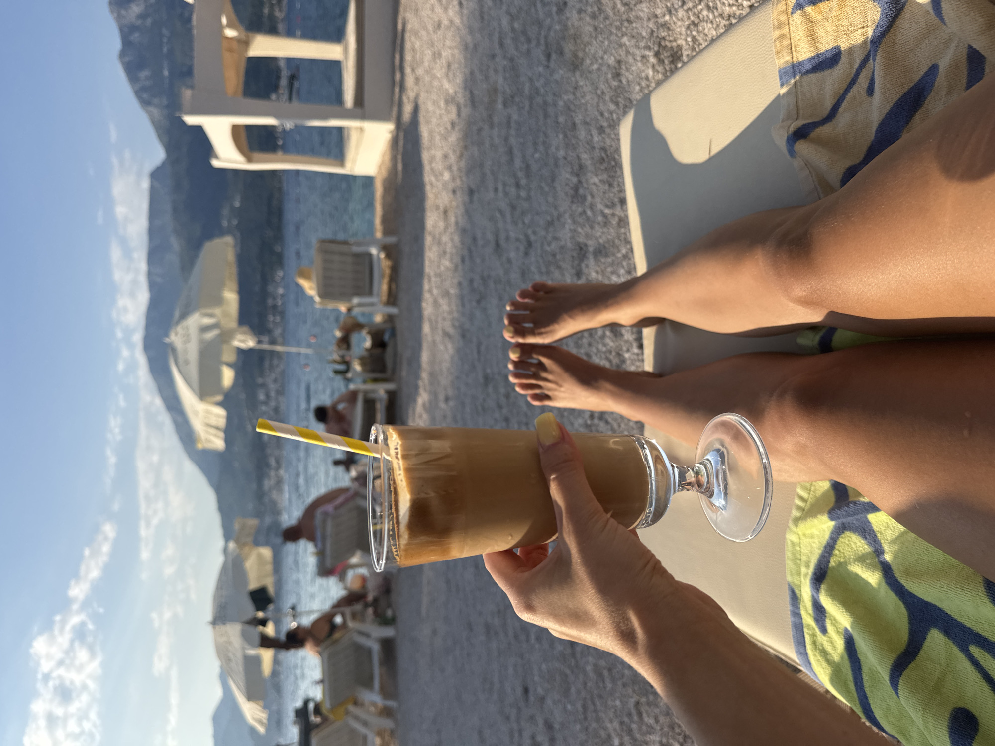 In a close-up, POV shot, a person's hand holds a tall glass of iced coffee with a yellow and white striped straw. The drink is positioned in the foreground, with the person's legs and feet visible below, resting on a lounge chair covered with a yellow and blue patterned towel. The background depicts a sunny beach scene with a calm sea, mountains in the distance, several beach umbrellas, and other beachgoers lounging on chairs. The lighting suggests it is late afternoon or early evening.