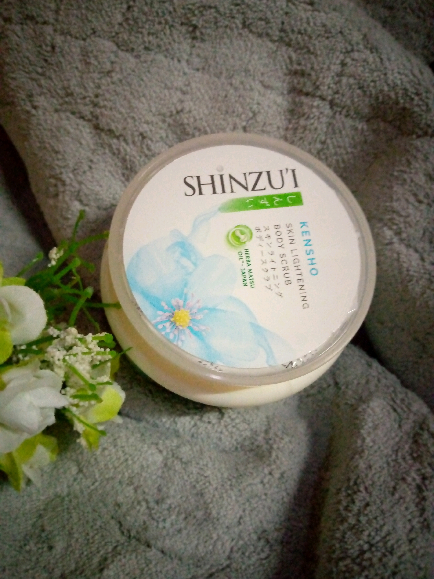The image shows a round, clear plastic container of Shinz'ui Kensho Skin Lightening Body Scrub on a textured gray fabric surface. The container has a white label with blue and green accents. The label features text in English and Japanese, and a watercolor illustration of a blue flower with a yellow center. A small sprig of artificial white flowers and green foliage is positioned to the left of the container. The fabric appears to be a towel or blanket. The scene does not contain any specific identifiable landmarks or features that would indicate a location in Sumberwono, Indonesia.