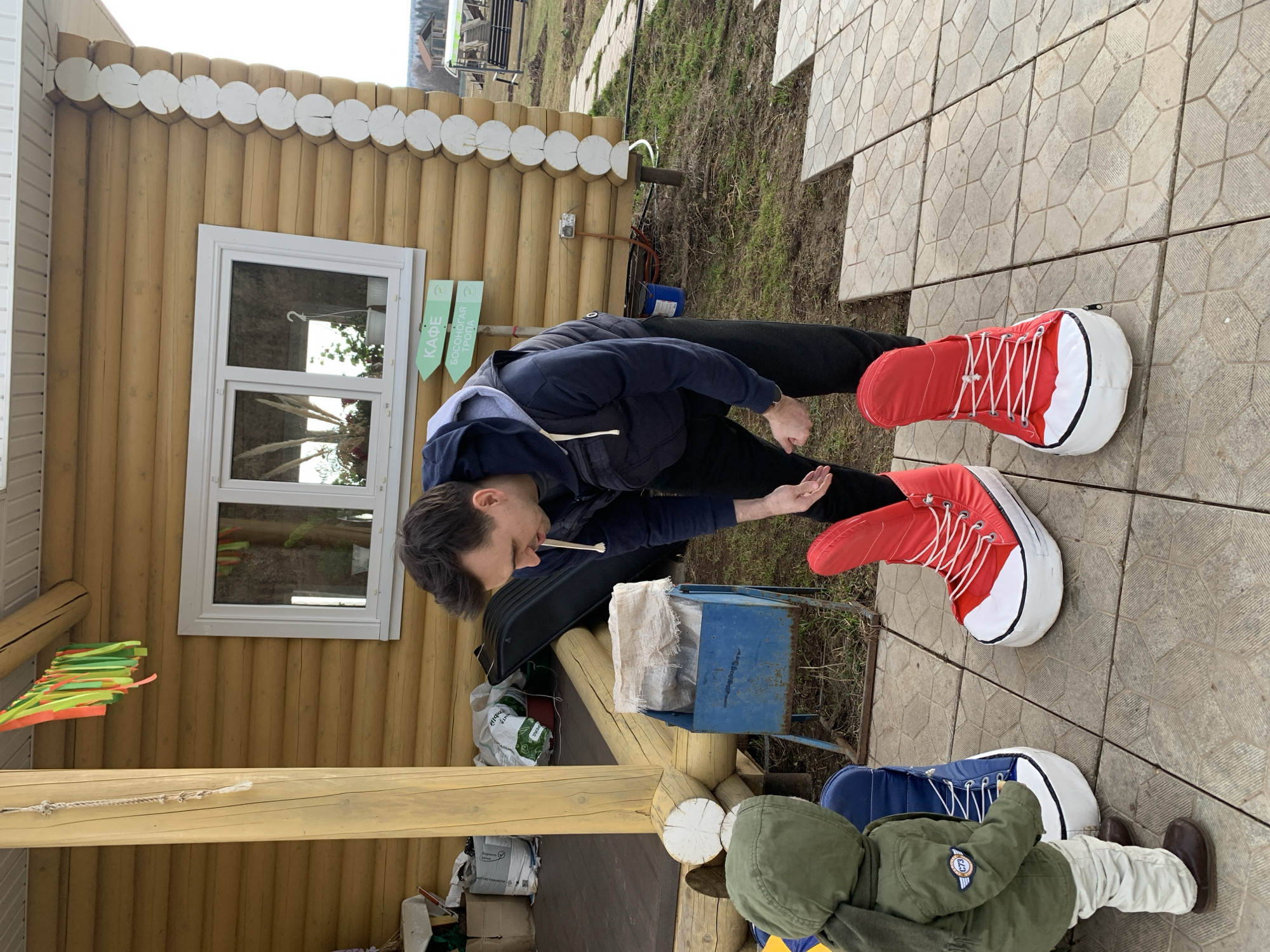 In Sepych, Russia, a man stands outdoors wearing oversized red and white sneaker costumes, bending over a blue metal box, possibly examining or using it. He is dressed in a dark jacket and pants. To his right, a child is sitting with their back facing the camera, clad in an olive green jacket and beige pants, with similarly oversized blue and white sneaker costumes on their feet. They are situated on a tiled surface. Behind the man is a wooden structure with a window and decorative elements, possibly a building or cabin. The environment includes vegetation and an overcast sky. Several bags and other items are visible near the building's base.