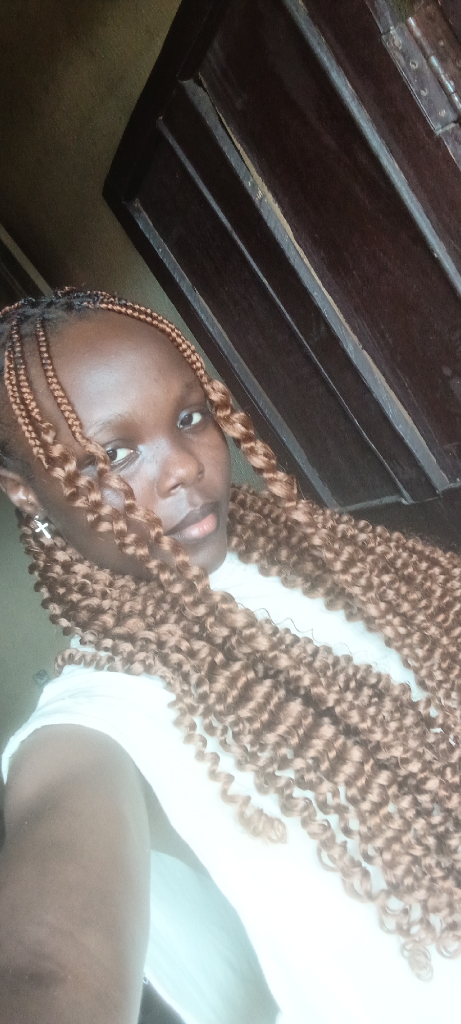 A close-up, slightly tilted portrait features a young woman with dark skin. Her hair is styled in long, voluminous, brown, curly braids that frame her face and extend down her chest. She wears a white top, parts of which are visible on her shoulders and chest. A silver cross earring hangs from her left ear. She looks directly towards the camera with a calm, neutral, or slightly pensive expression, her lips slightly parted. The background consists of a dark brown wooden door or paneled wall, featuring vertical lines and what appears to be part of a metal hinge on the right side. The lighting suggests an indoor setting, possibly during the daytime. The image was captured in Onitsha, Nigeria. No text is visible.