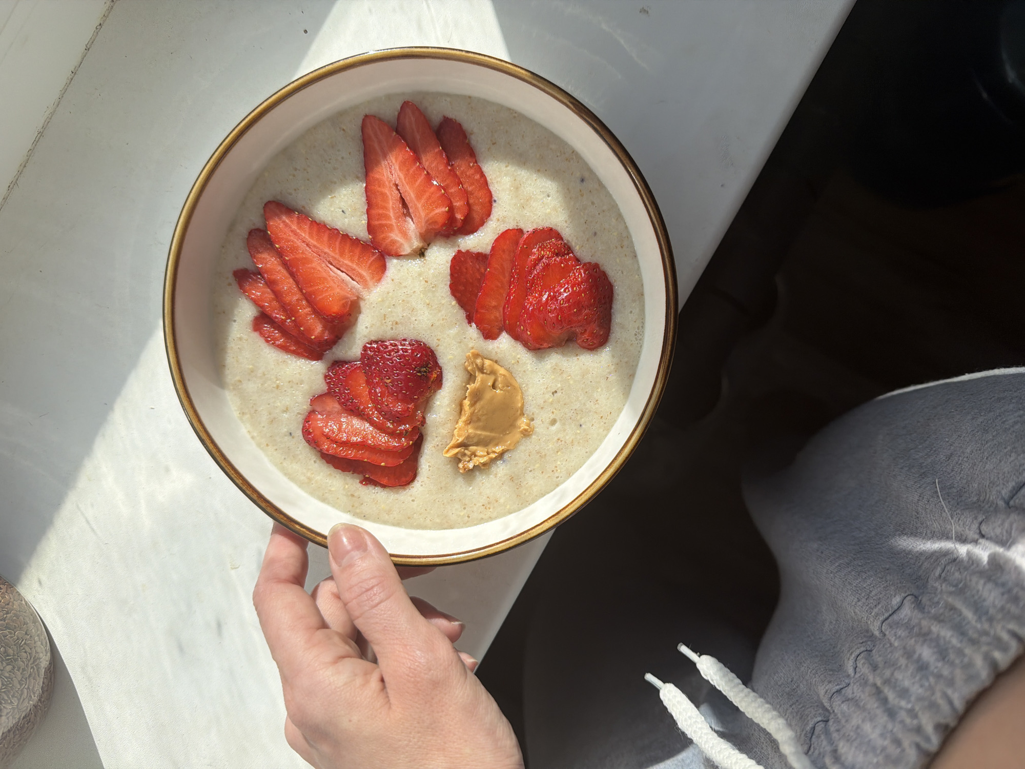 The image shows a person holding a bowl of oatmeal topped with sliced strawberries and a dollop of peanut butter. The bowl is cream-colored with a brown rim and is held in the person's left hand. The person is wearing gray sweatpants. The bowl is positioned near a window, with sunlight casting shadows on the white windowsill. The image does not contain enough information to determine the location as Perm, Russia.