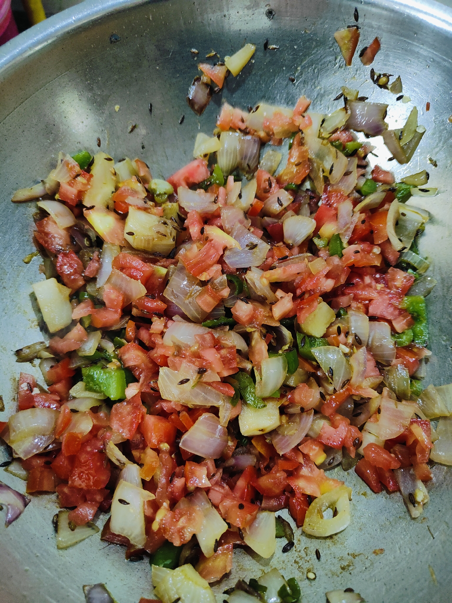 A close-up, overhead shot shows a metal pot filled with chopped ingredients for a dish, likely being prepared in a kitchen in Gurgaon, India. The ingredients include diced tomatoes, onions, and green chilies, mixed with whole cumin seeds. The metal pot appears to be slightly worn and used. The lighting suggests an indoor environment, possibly a home kitchen.