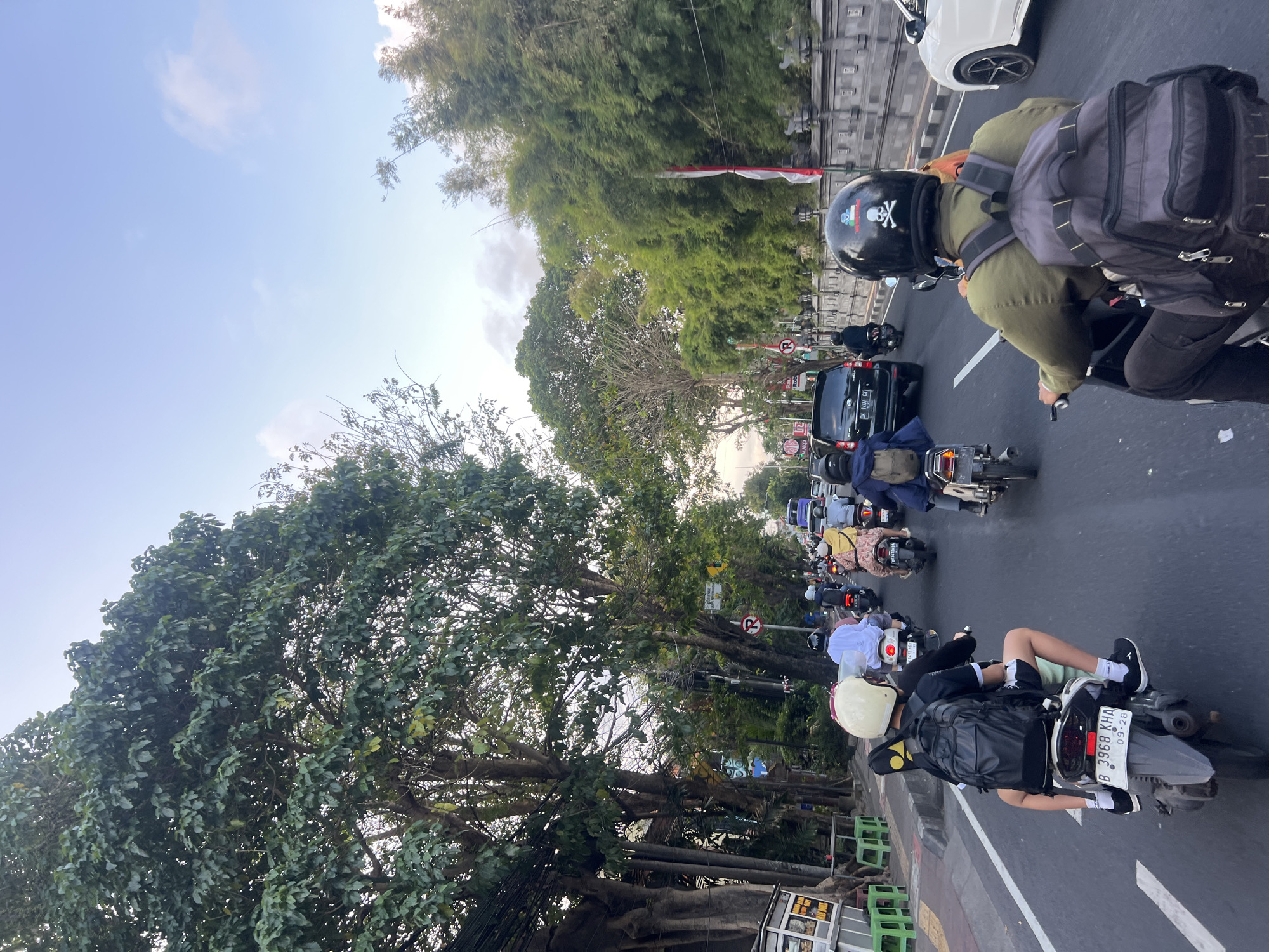 This is a first-person, wide-angle shot of a busy street in Denpasar, Indonesia, likely during late afternoon. The camera is positioned behind a rider on a motorcycle, looking forward. The street is congested with numerous motorcycles and a few cars.
In the foreground, the motorcycle rider being filmed wears a dark green jacket and a black helmet adorned with a skull and crossbones emblem. They are carrying a large, dark grey backpack. The motorcycle's license plate is visible and reads "DK 1407 GI". To the left of the motorcycle, another rider with a white helmet and a black backpack is partially visible.
The road itself is paved with asphalt and marked with white lines. On either side of the street, lush green trees with dense foliage line the way, casting shadows. Some of the trees are very large and mature, with thick trunks. Beyond the trees, there are glimpses of buildings and more vegetation, including bamboo. A traditional stone wall with decorative elements is visible on the right side of the road, with Indonesian flags affixed to poles.
Traffic signs are present, including a red circle with a horizontal bar indicating "No Entry" and a blue circular sign. A black car is visible in the middle distance, and further ahead, a white car is moving in the opposite direction. The sky is a pale blue with a few wispy clouds, suggesting it might be nearing sunset. The overall atmosphere is one of a typical urban traffic scene in a tropical environment.