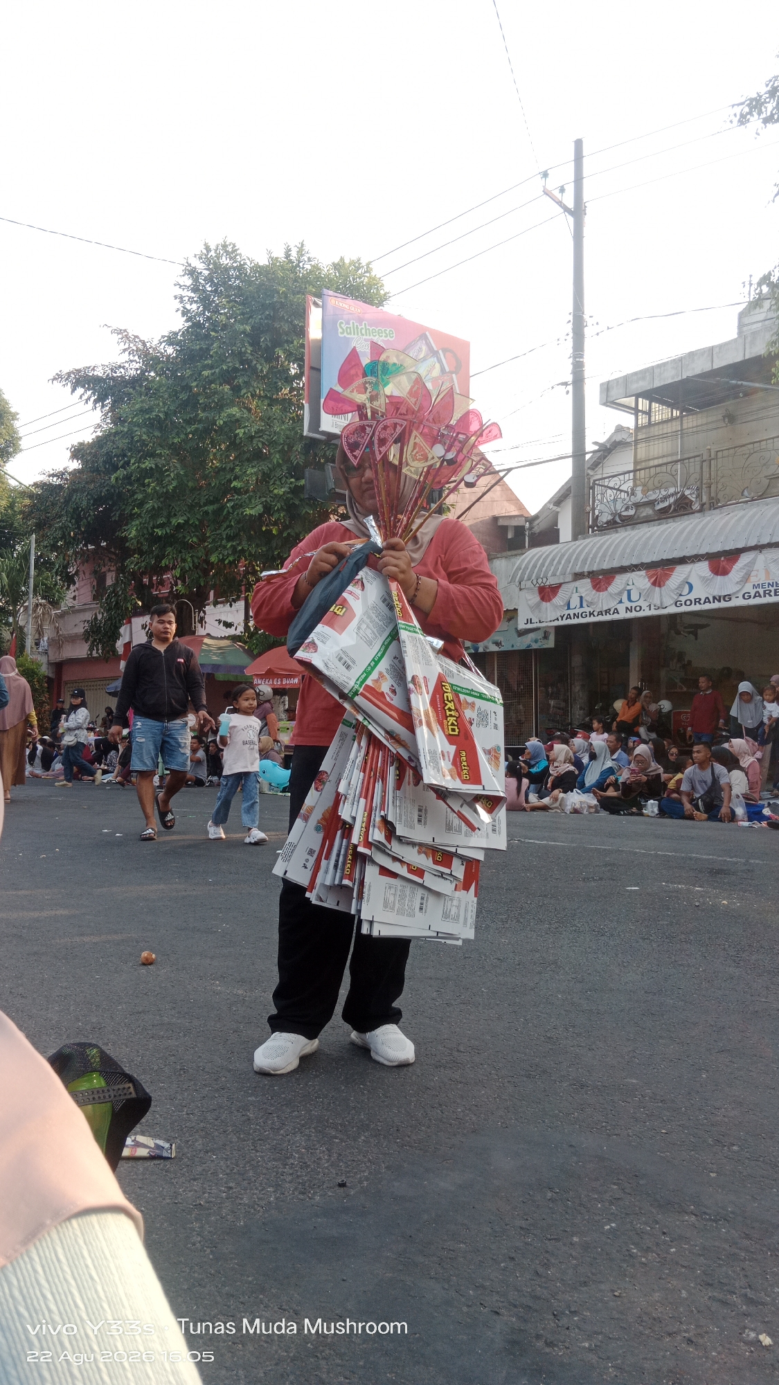 In the city of Genengan, Indonesia, a vendor displays a collection of pinwheels and packaged goods for sale. The vendor, wearing a red shirt and black pants, holds a bundle of colorful pinwheels above their head and several large, white packages in their arms. The packages appear to be food items, with visible branding. In the background, a billboard advertises "Salticheese" and other products. A crowd of people is gathered, some sitting on the ground, suggesting a lively street market or community event. The scene is set on a paved road, with trees and buildings lining the sides, indicating an urban or suburban environment. The time of day appears to be late afternoon, with long shadows cast by the setting sun.