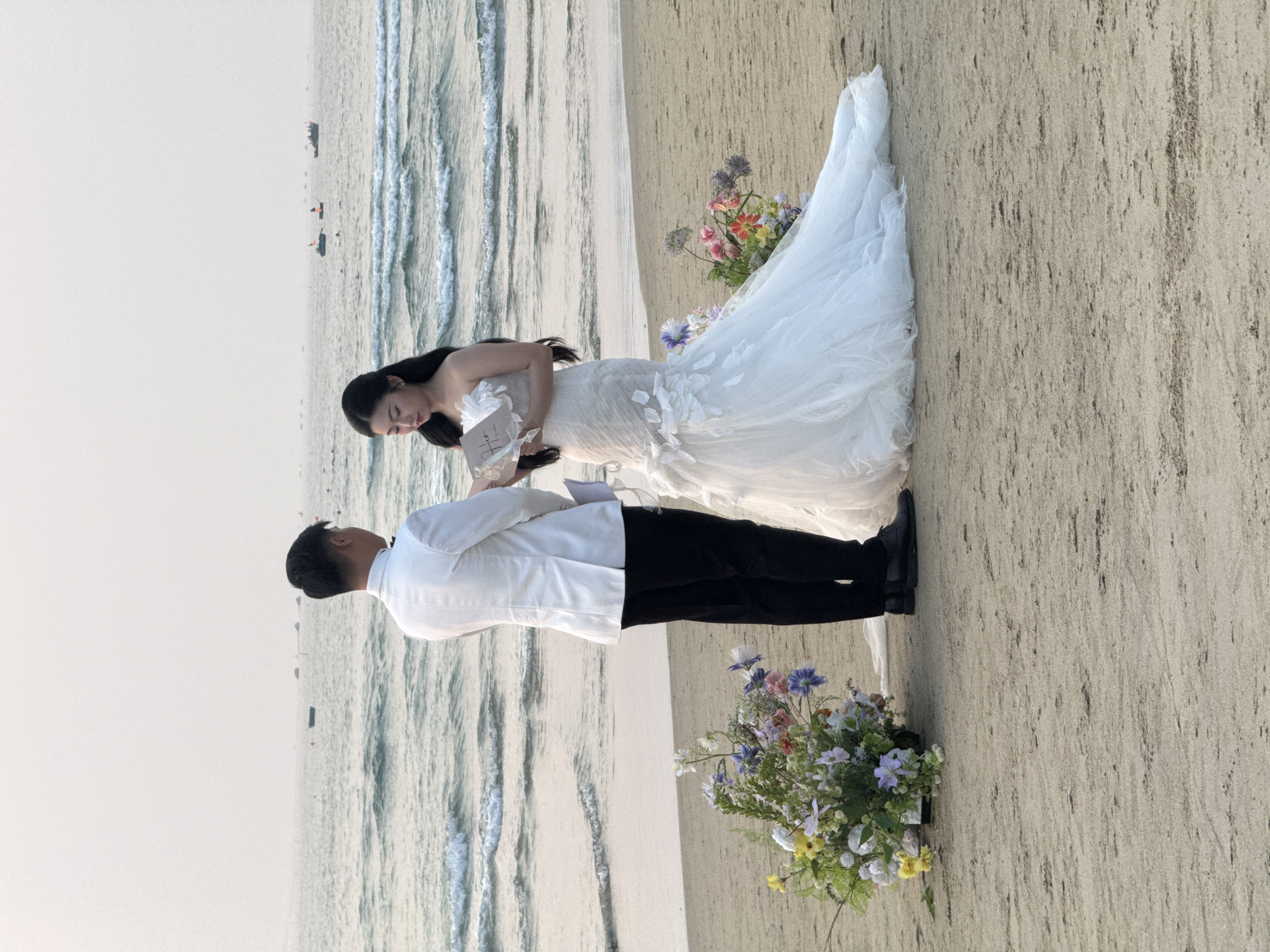 The image shows a wedding ceremony taking place on a beach. The bride, wearing a white wedding dress, is standing opposite the groom, who is wearing a white shirt and black pants. Both are holding cards. There are flower arrangements on the beach near them. The beach is bordered by the ocean. The sky is overcast. Based on the ocean and beach characteristics, this image could be of a wedding ceremony taking place on a beach in the city of Đà Nẵng, Vietnam.