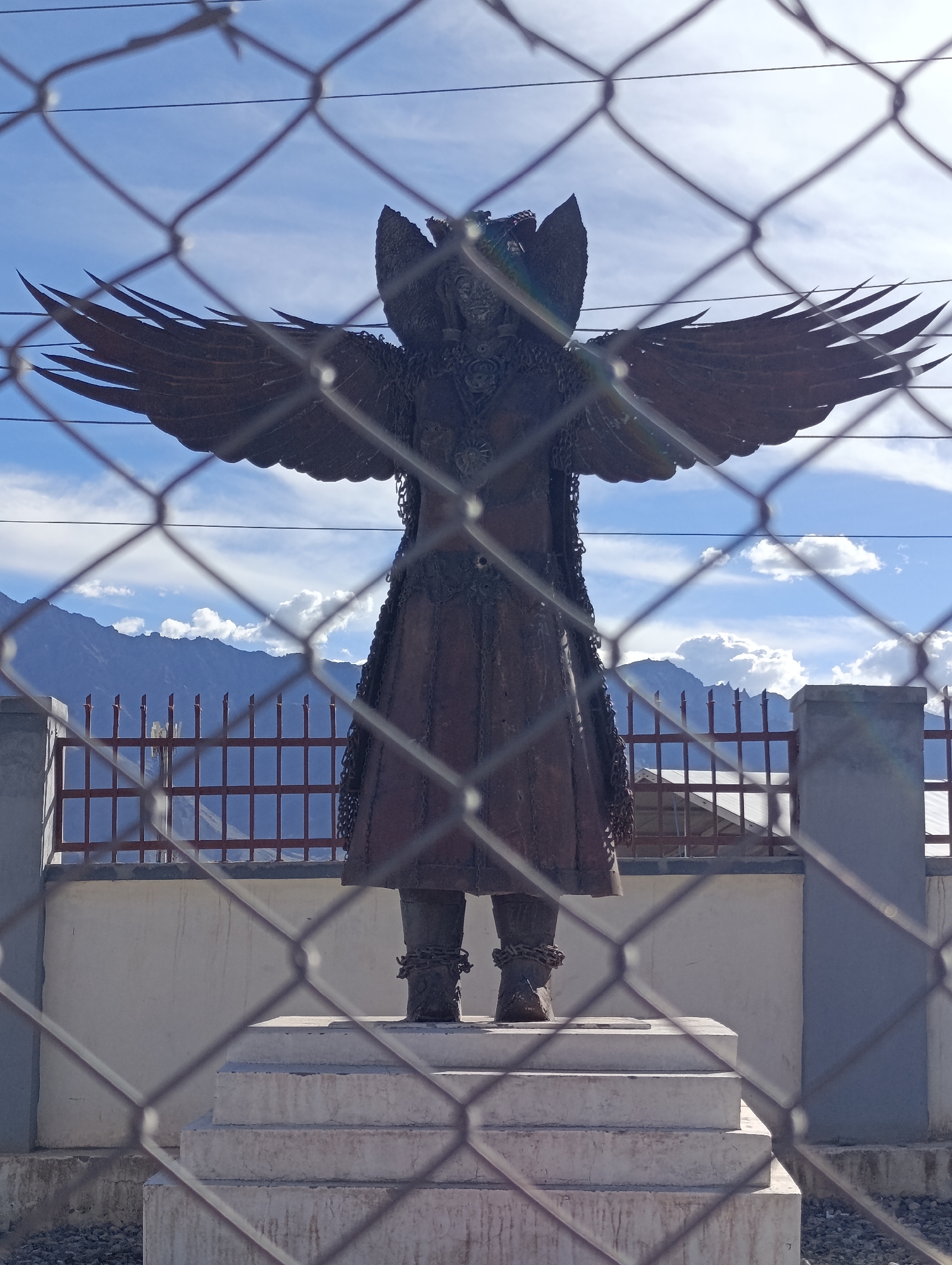 A full-body, eye-level shot captures a dark, weathered statue of a winged humanoid figure behind a chain-link fence. The figure stands erect, with arms outstretched to the sides, showcasing large, feathered wings. The statue is adorned with a segmented skirt and has what appear to be chains or ropes wrapped around its ankles. It rests on a multi-tiered concrete pedestal. The background reveals a rugged mountain range under a partly cloudy blue sky. A fence with a red horizontal bar is visible in front of the mountains, and a few buildings with tin roofs are situated beyond it. The foreground is dominated by the diamond pattern of the chain-link fence, obscuring parts of the statue and its immediate surroundings. The image is taken outdoors in a daylight setting. The statue is located at the Hall of Fame Museum in Leh, India, which is a museum showcasing the history of the Indian Army in the region and the experiences of soldiers in the Himalayas.