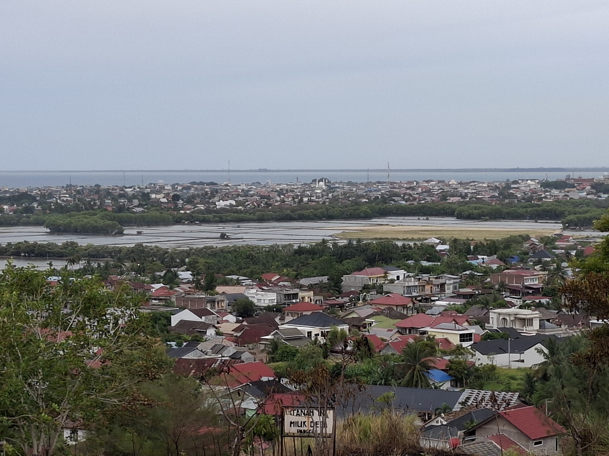 This is an elevated daytime view of a portion of the city of Lhokseumawe, Indonesia. In the foreground, there are several residential houses with red and gray roofs, interspersed with trees and shrubbery. A sign partially obscured by foliage is visible, with the words "TANAH MILIK" visible. Beyond the residential area, there's a strip of land featuring a lagoon or wetland, bordered by trees. In the distance, there's a densely populated area with a mix of low-rise buildings, stretching to the coastline where the water meets the sky under an overcast sky. Tall antennas are visible on the horizon. The photo appears to have been taken from a higher vantage point, possibly a hill or elevated structure.