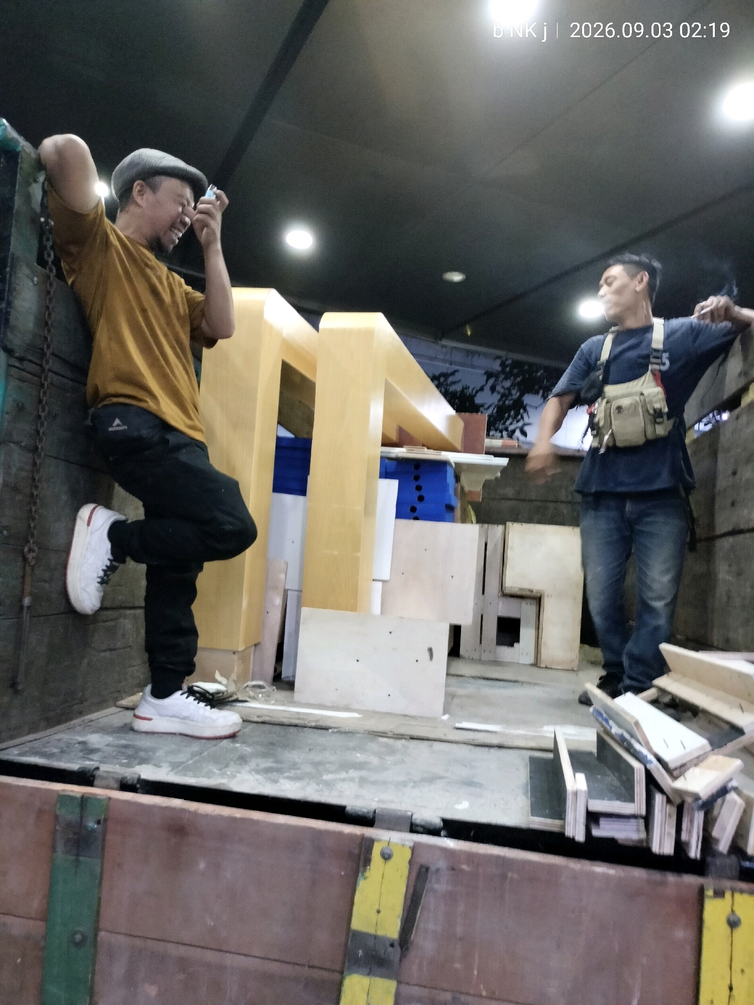 Two adult males are inside a truck bed. One male, on the left, is wearing a mustard yellow t-shirt, dark pants, a grey flat cap, and white sneakers. He is leaning against the truck wall and holding a small object up to his eye, possibly a camera or phone. He appears to be laughing or smiling. The other male, on the right, is wearing a dark blue t-shirt with a tan utility vest, blue jeans, and dark shoes. He is smoking and looking to his left. The truck bed is filled with various wooden materials and some blue plastic crates. The scene is lit by overhead lights. The date stamp on the image reads "b NK j | 2026.09.03 02:19". The image is taken in South Jakarta, Indonesia.