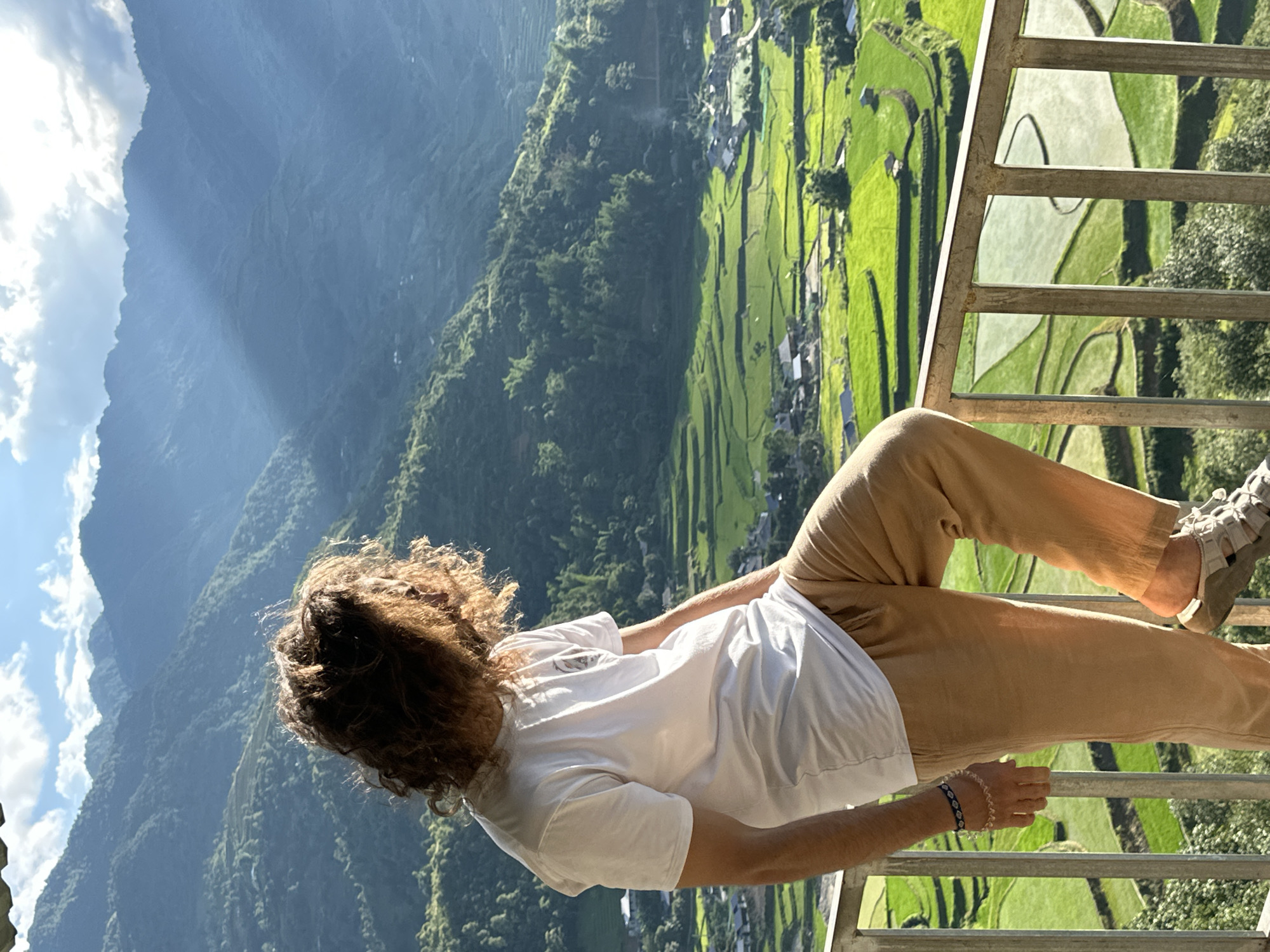 A young adult male, seen from the waist up and turned away, stands on a metal balcony overlooking a mountainous landscape in Tú Lệ, Vietnam. The individual has long, curly brown hair, significantly backlit by the sun, creating a bright halo effect around the head. He wears a white short-sleeved t-shirt, light brown or khaki long pants, and light-colored sandals. Two bracelets, one dark and beaded, the other lighter and possibly woven, are visible on his left wrist. His right foot is propped on the lower bar of the light grey metal railing, and his head is turned towards the right, gazing at the distant scenery.
Below the balcony, vibrant green terraced rice fields stretch across the valley floor, interspersed with small residential structures. Some sections of the rice fields appear flooded, reflecting light. Beyond the fields, multiple layers of lush green mountains rise, fading into a hazy blue-grey distance under a partially cloudy sky. Sunlight breaks through the clouds on the upper right, casting visible rays onto the distant mountains. The overall lighting suggests either early morning or late afternoon.