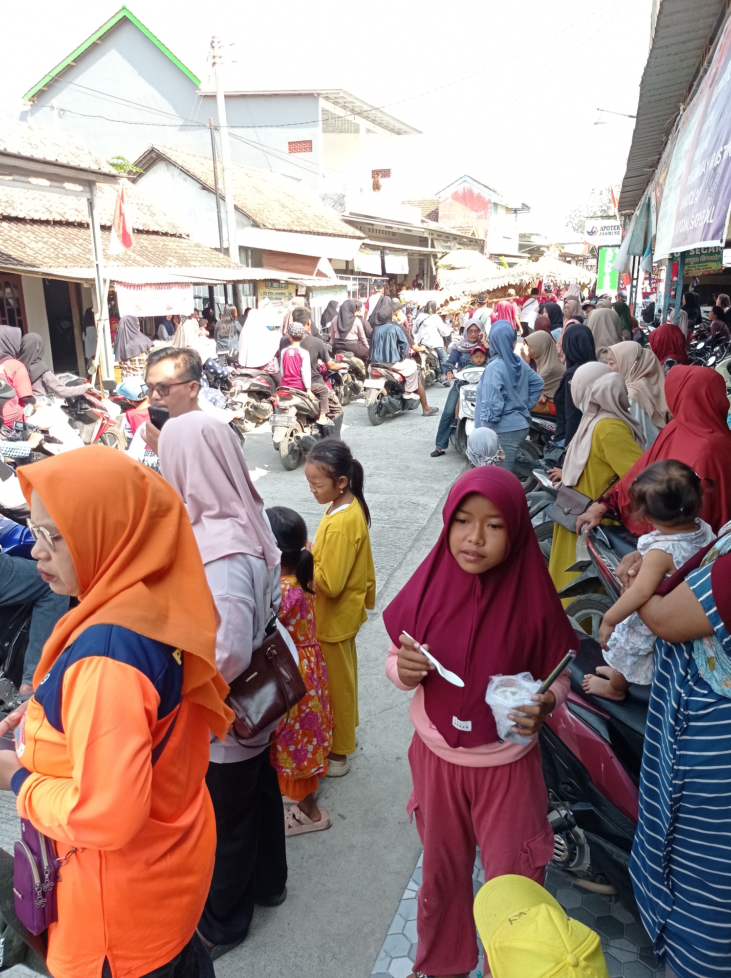 In the city of Maribaya, Indonesia, a crowded street scene unfolds under a bright sky. The image captures a bustling marketplace or thoroughfare lined with stalls and shops. People, mostly women and children, are seen navigating the area, many wearing hijabs and traditional attire. Motorcycles are a prominent mode of transportation, parked and utilized by individuals throughout the scene.
In the foreground, a young girl in a maroon hijab is holding a spoon and a cup of what appears to be ice cream. Beside her, an adult woman in an orange hijab is partially visible, facing left. Behind them, a child in a yellow outfit stands, while other individuals are seen in the background engaged in various activities.
The environment suggests an outdoor market or a busy street with buildings on either side. The architecture of the buildings appears to be a mix of traditional and modern styles. Signs and banners are visible, indicating commercial establishments. The overall atmosphere is one of lively activity and daily life in a community setting.