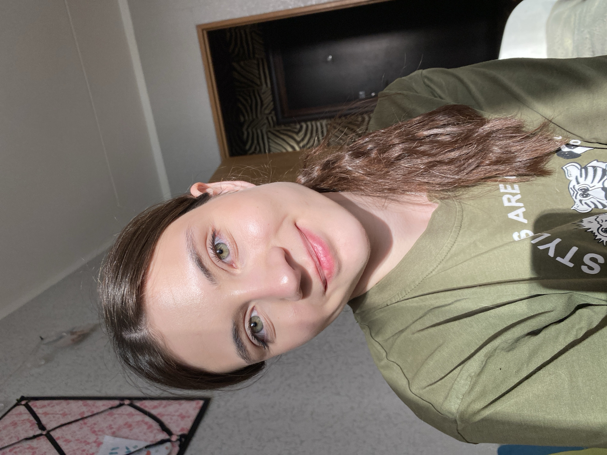 A selfie of a woman with olive-colored eyes and long, brown hair, wearing a green t-shirt with white text and a tiger graphic. The background includes a wall and a partially visible headboard with a decorative design. Based on the provided information, the location is Votkinsk, Russia, though no specific location within the city can be determined from the image.