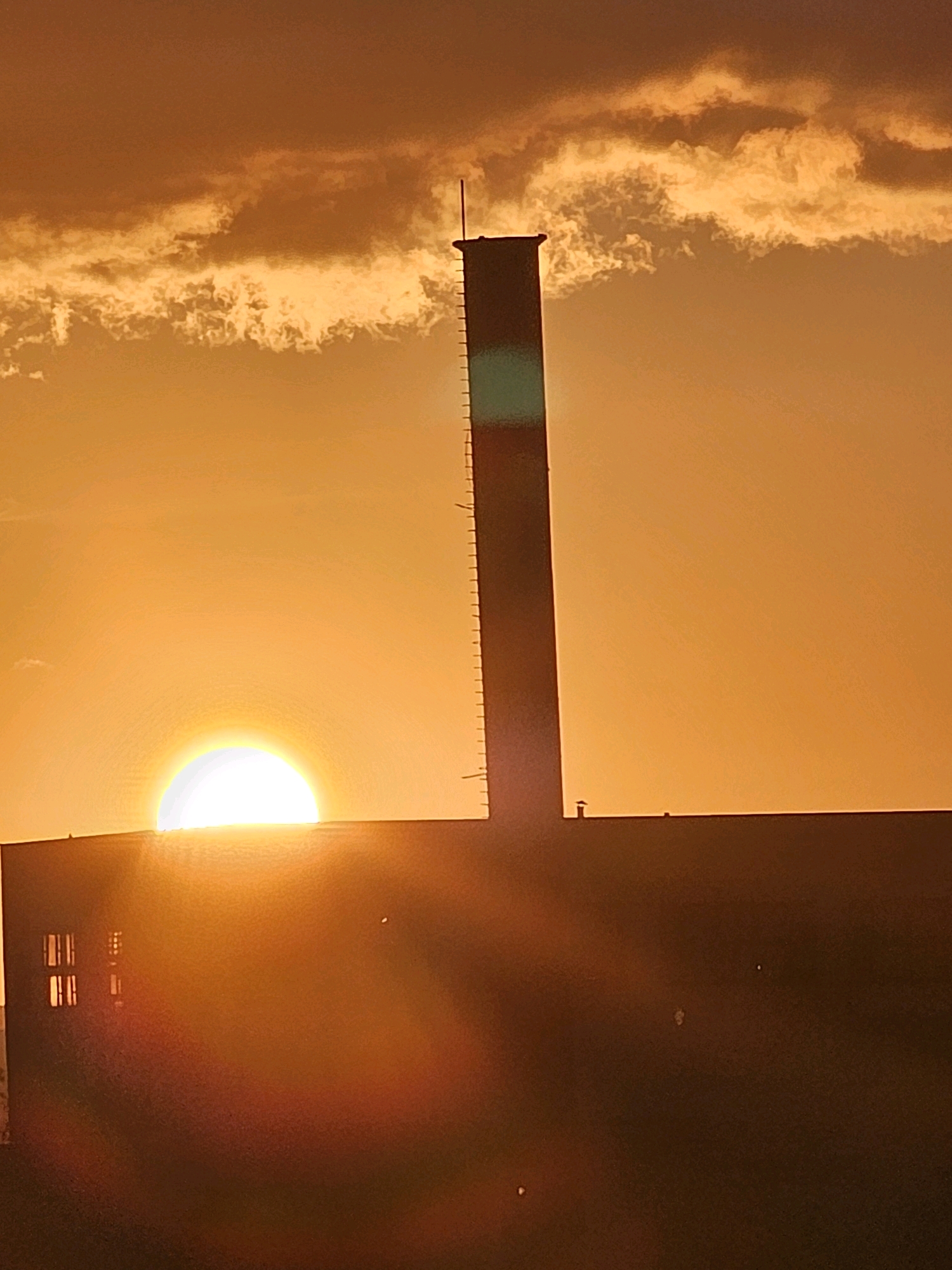 This is a photograph of a sunset over a building with a tall, dark smokestack. The sun, a bright white disk, is partially obscured by the top of the building and the smokestack. The sky is a vibrant orange, with wispy clouds that are illuminated by the setting sun. The building appears to be industrial, with several windows visible on its facade. The smokestack is rectangular in shape and has a series of horizontal lines etched into its surface. There are no people visible in the image. The scene is set in the city of Khimki, Russia.
