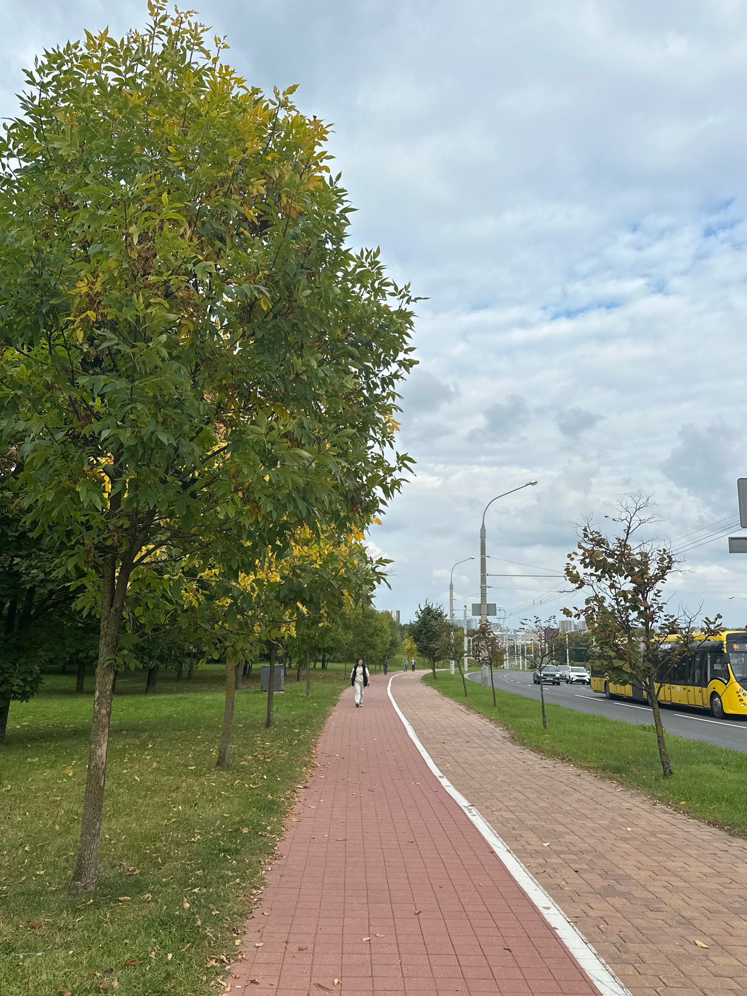 The image captures an outdoor scene along a paved pathway, bordered by a grassy area on the left and a road with traffic on the right. A person is walking away from the camera on the pathway. The pathway is made of red bricks and has a white line marking its edge. Trees line the grassy area, some of which show signs of autumn with yellowing leaves. On the road, a yellow bus and several cars are visible. The sky is overcast with clouds. This scene is consistent with an urban park or a wide boulevard in Minsk, Belarus, given the presence of public transport like the yellow bus and the general urban infrastructure.