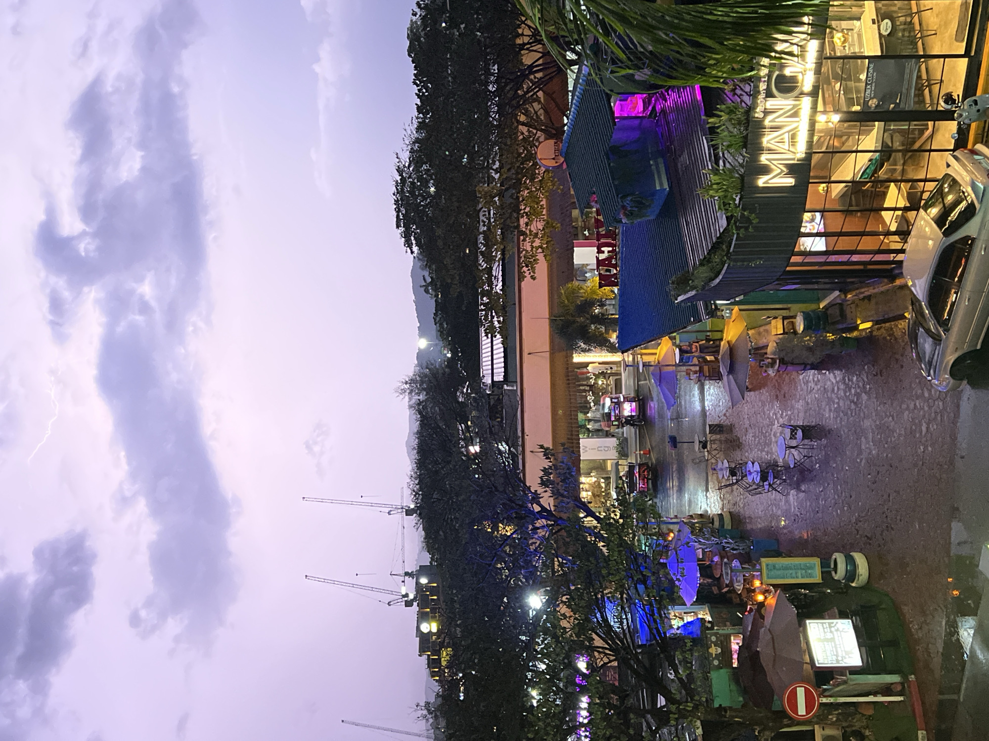 The image captures a street scene in an urban setting, possibly during or after rainfall, given the wet appearance of the ground. The composition includes elements of both natural and built environments.
To the right, there is a restaurant or establishment named "MANO" with illuminated signage, indicating it might be evening or night. There is a car parked nearby. Further down the street, there are outdoor seating areas with tables and chairs, some possibly under umbrellas, suggesting dining or leisure activities.
On the left side of the image, the sky is visible, presenting a cloudy or stormy atmosphere. There is what appears to be a lightning strike visible in the sky. Several construction cranes are silhouetted against the backdrop of the sky. The presence of trees and other foliage suggests some level of landscaping or vegetation in the area.
Overall, the image portrays a street view with commercial establishments, outdoor seating, and a slightly dramatic sky featuring lightning and construction cranes. The lack of people in clear view suggests a moment of quiet or sparse activity in the urban environment.