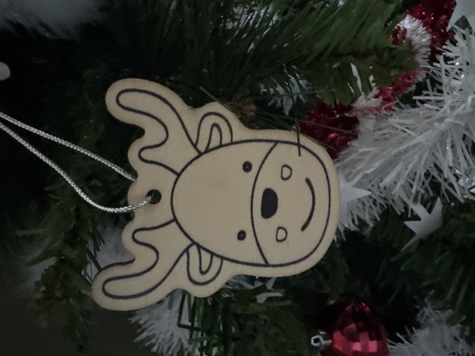 A close-up image captures a light brown, flat, reindeer-shaped ornament with black outlines defining its face, including two black dot eyes, a larger black oval nose, a smiling mouth, and stylized antlers. A thin, silver-colored string forms a loop through a small hole at the top of the ornament, suspending it. The ornament hangs on an artificial Christmas tree, characterized by dark green pine needles visible in the background. Other decorations partially observed include a shiny red spherical ornament in the lower-left foreground and segments of white and red tinsel or garland in the lower-right background. The scene is located in the city of Le Puy-en-Velay, France.