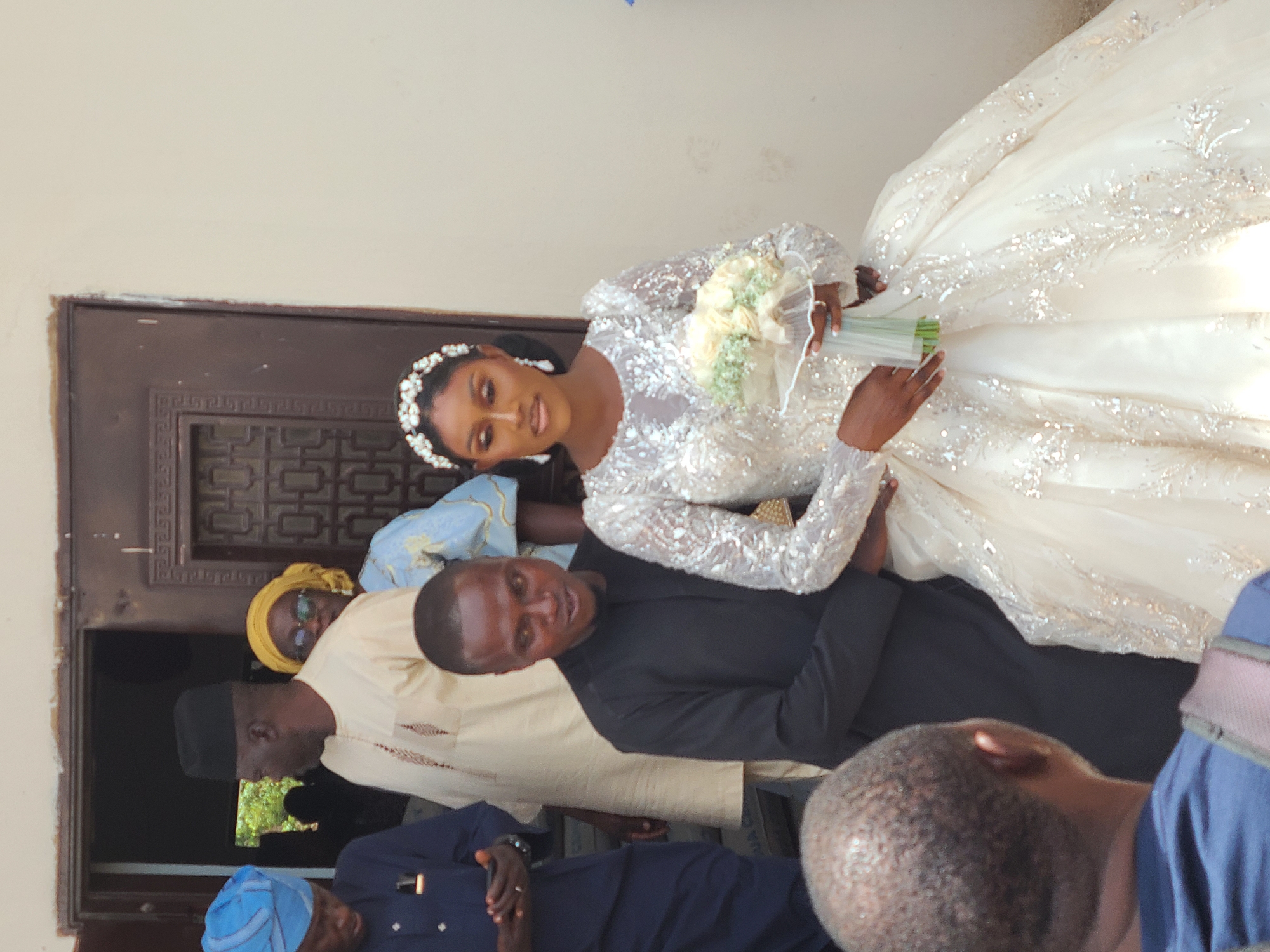 The image depicts a wedding scene. A bride in a white, long-sleeved gown, adorned with glitter, holds a bouquet of white flowers. She stands next to a man in a black shirt. Several other people are present, including an individual in a blue outfit, another in a tan garment, and a woman wearing a yellow headscarf. The scene appears to be taking place in front of a building with a decorated door. It's in the city of Barakin Lerawi, Nigeria.