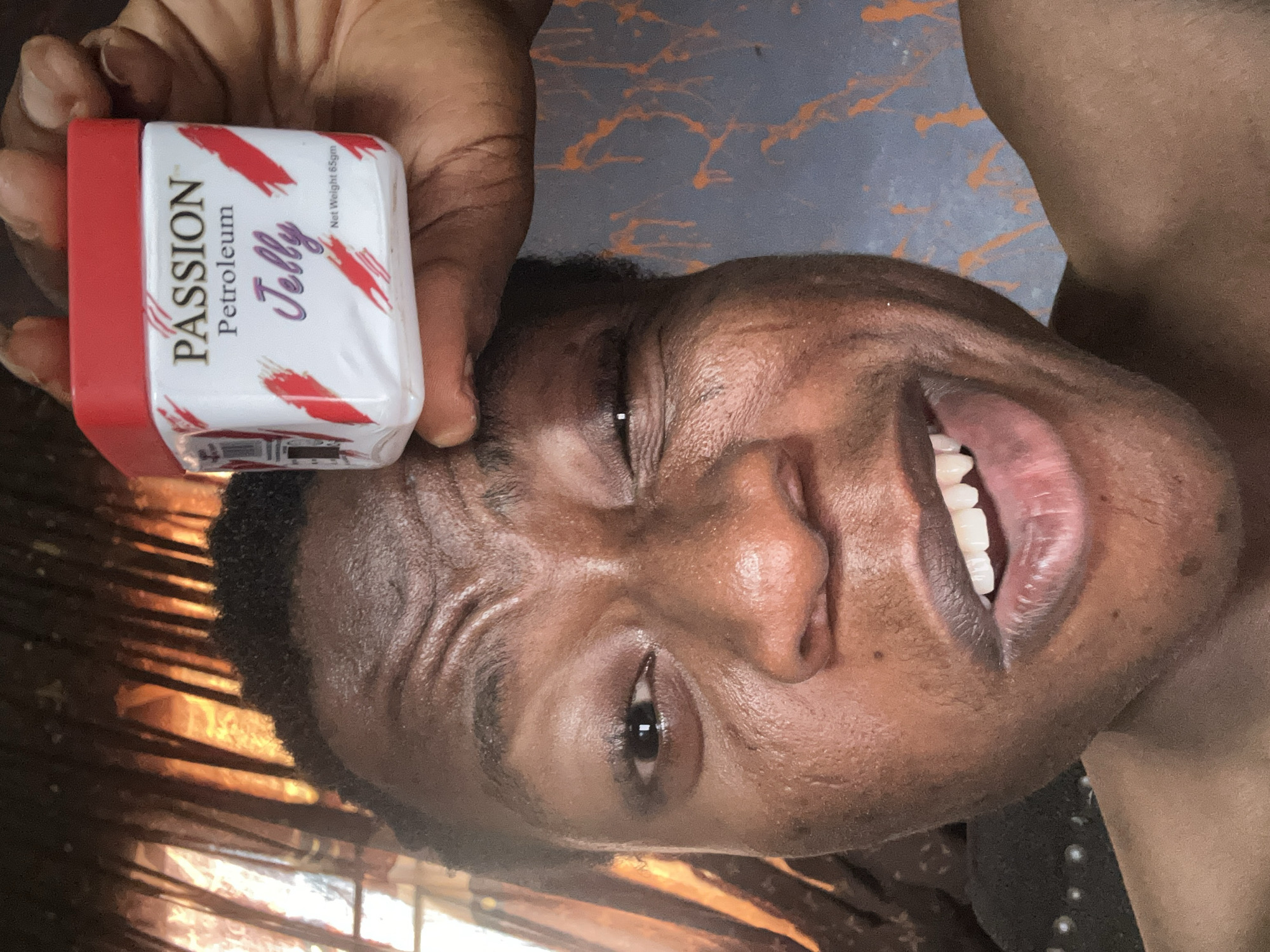 The image shows a person with dark skin holding a container of "Passion Petroleum Jelly" next to their face. The container is white with a red lid and has red brushstroke designs. The person is smiling and looking towards the camera. The background is blurry and indistinct. The location is Unknown, Unknown.