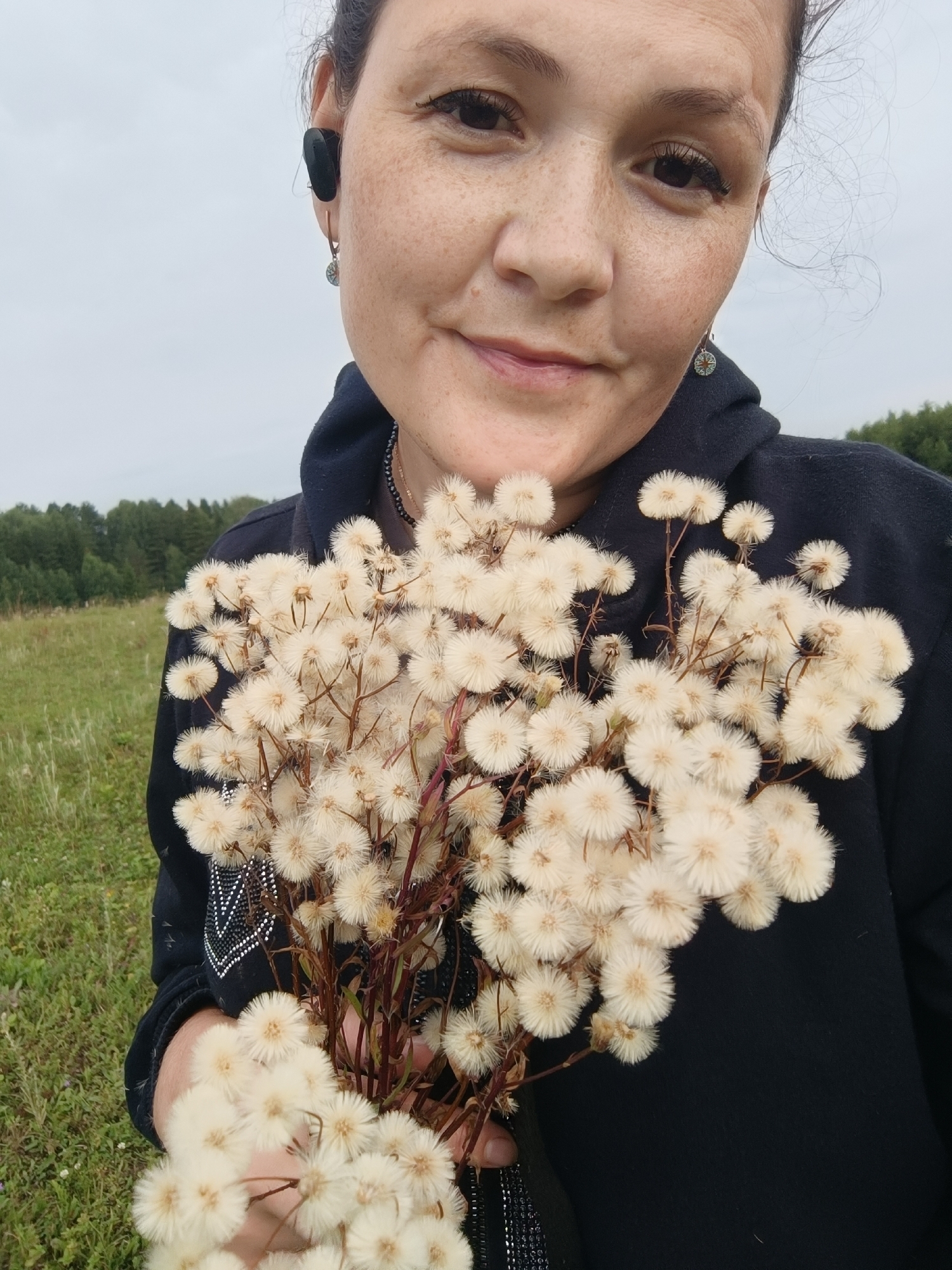 A close-up medium shot captures a woman from the chest up, holding a bouquet of fluffy white wildflowers. She has dark hair pulled back, a light complexion with freckles, and is wearing a dark hooded sweatshirt. A black earbud is visible in her right ear, and she wears a small circular earring in her left ear. The background consists of a grassy field with a line of trees in the distance under an overcast sky. The image does not provide any information about the location within Karsha, Russia.