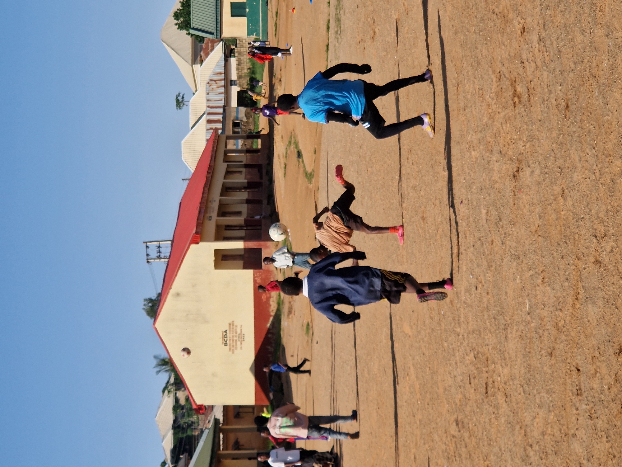 This image depicts a group of boys playing football on a dusty, outdoor field in Kabong, Nigeria. The field is a sandy, brown expanse with sparse vegetation and some scattered debris. In the background, there are several low-rise buildings with red and yellow exteriors, typical of the region. Some buildings have corrugated metal roofs, and one has "BCDA" signage. A clear blue sky stretches above the scene.
Several boys are actively engaged in the football game. Two boys in the foreground are central to the action: one in a blue shirt and black pants is poised to kick the ball, while another in a brown shirt and dark shorts is mid-air, attempting to kick the ball as well. Other boys are visible in the background, some running, others observing the game, and one appears to be kicking the ball toward the center of the field. The boys are dressed in casual attire, including t-shirts, shorts, and athletic shoes. The atmosphere appears energetic and playful, indicative of a spontaneous game among the local youth.