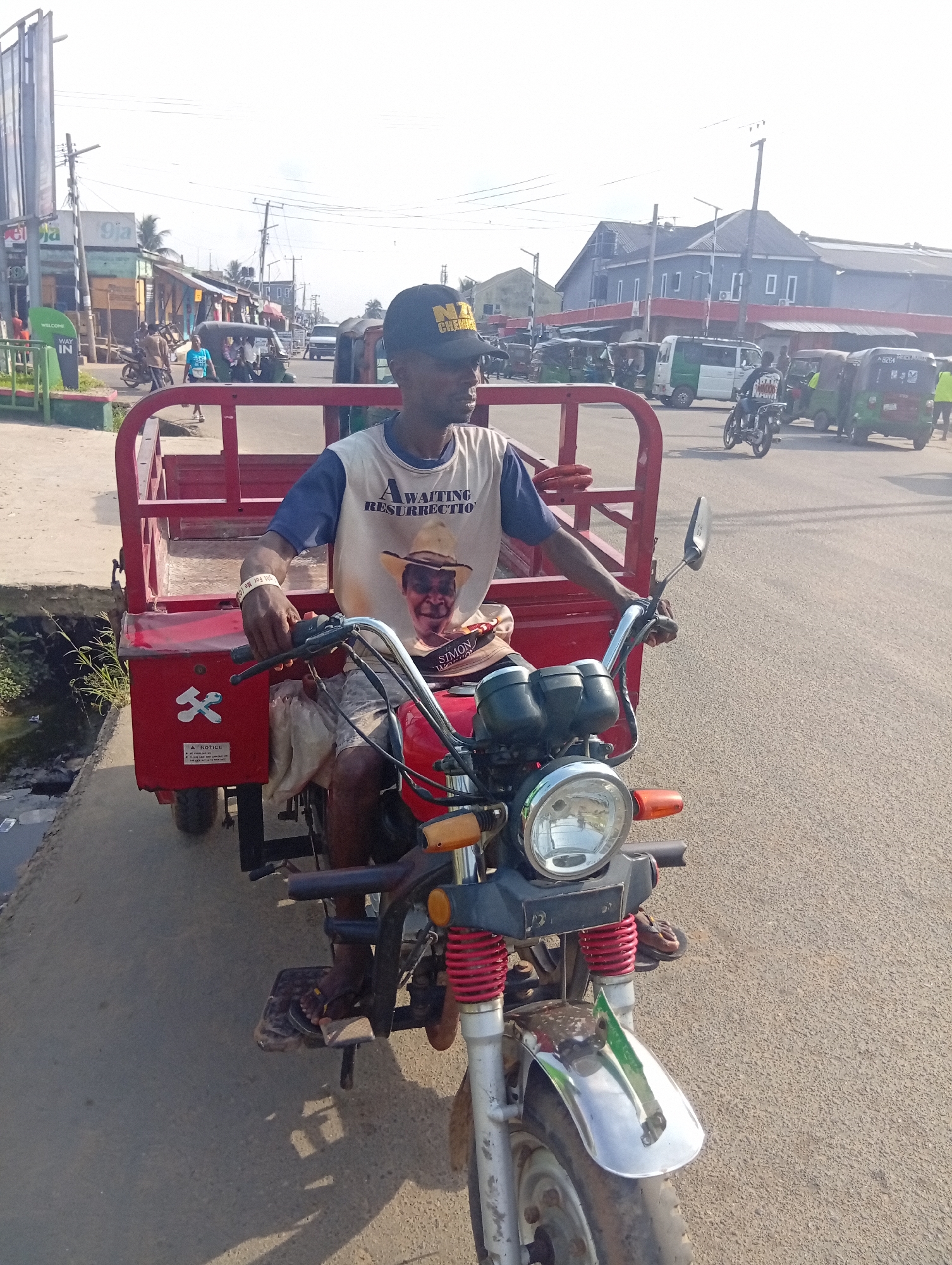 The image shows a man on a red three-wheeled motorcycle, known locally as a "Keke" or "Tricycle", in an urban setting. The man is wearing a black cap with yellow writing that appears to say "NA CHEMIST", a T-shirt with a picture of a man wearing a hat and the words "Awaiting Resurrection" written above it, and he has a white wristband. He is looking to his right. The Keke has a red cargo bed behind the driver's seat.
In the background, there is a busy street scene with other Keke's, motorcycles, and vans. There are buildings visible, some of which appear to be shops or businesses. Utility poles and wires are also visible. The image appears to be taken on a sunny day.
The location is likely Aba, Nigeria, based on the presence of the Keke tricycle taxis, which are common forms of transportation in many Nigerian cities, including Aba.