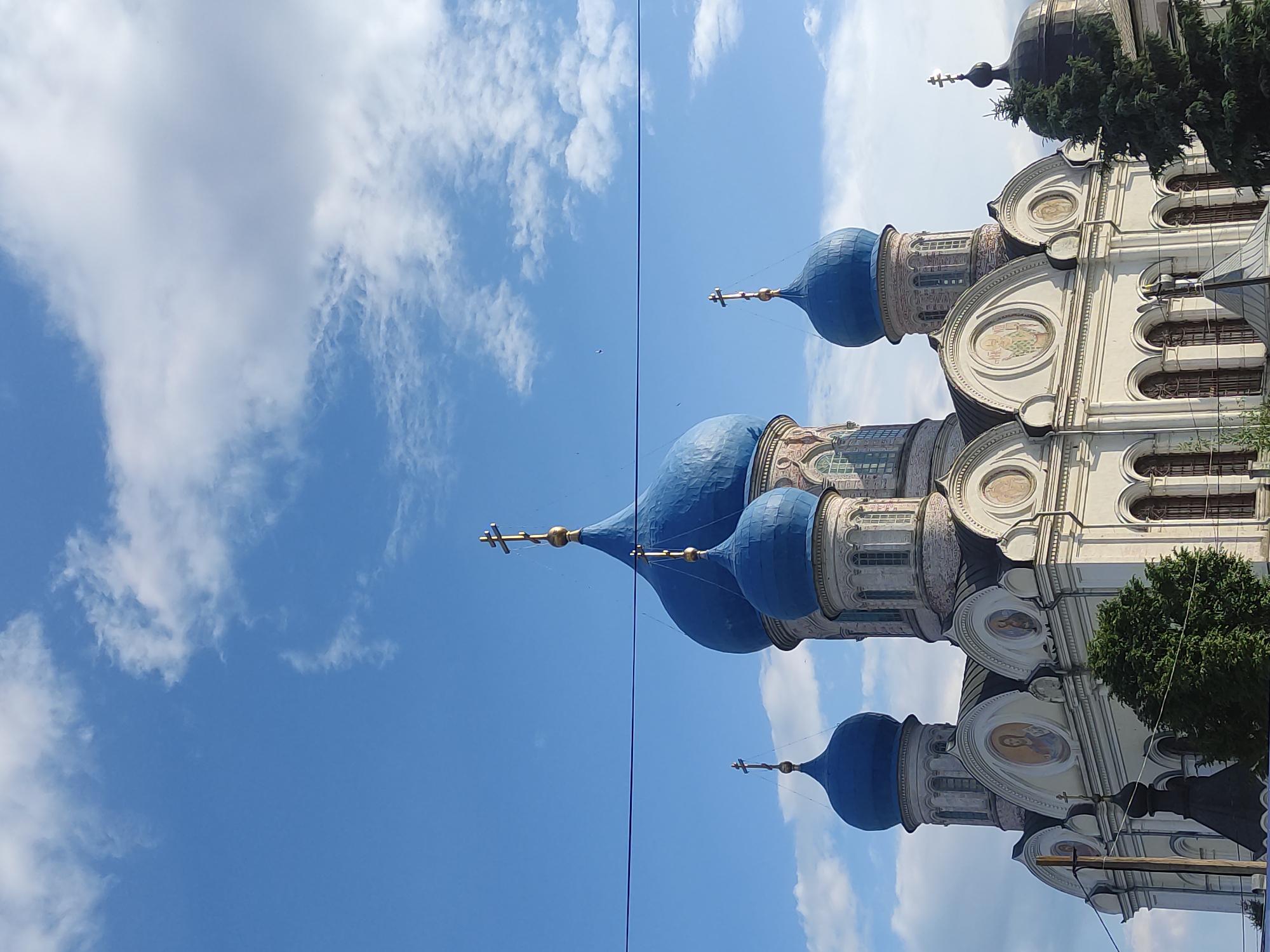 This image shows a medium shot of the Church of the Transfiguration, a domed Orthodox church located in Rogachovo, Russia. The church has a white facade with several blue domes topped with golden crosses. The main dome is the largest, with smaller domes surrounding it. The church features arched windows and decorative elements, including circular paintings on its facade. In the foreground, a power line runs horizontally across the frame. Green trees are visible to the right and left of the church, and a dark, smaller dome structure is partially visible on the right. The sky above is blue with scattered white clouds. No people or actions are depicted in the image.