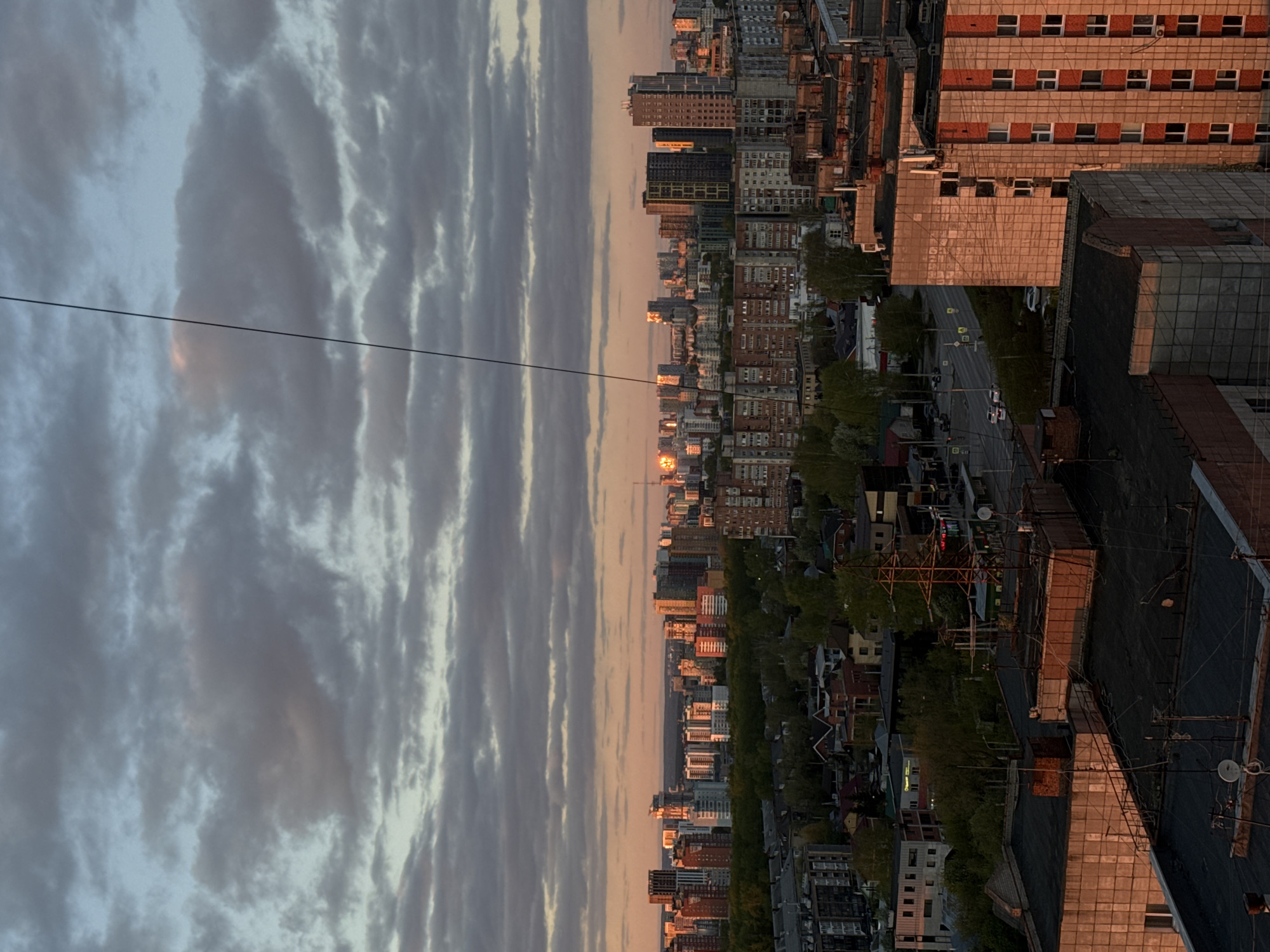 The image depicts a sunset view of Perm, Russia. The photograph is taken from an elevated position, offering a panoramic vista of the cityscape, partially obscured by foreground architecture. The sky is filled with striated altocumulus clouds. The setting sun casts a golden hue on the cityscape. Buildings of varying heights and architectural styles stretch into the distance. A powerline cuts across the upper portion of the sky. The architecture in the foreground appears to be industrial or residential buildings, characterized by flat roofs and simple construction. The foreground structures cast long shadows, indicating that the sun is low on the horizon. A network of streets and roads interspersed with trees is visible in the midground, with vehicles plying the routes. The presence of multiple windows and other architectural details suggests that the buildings are inhabited or in use.