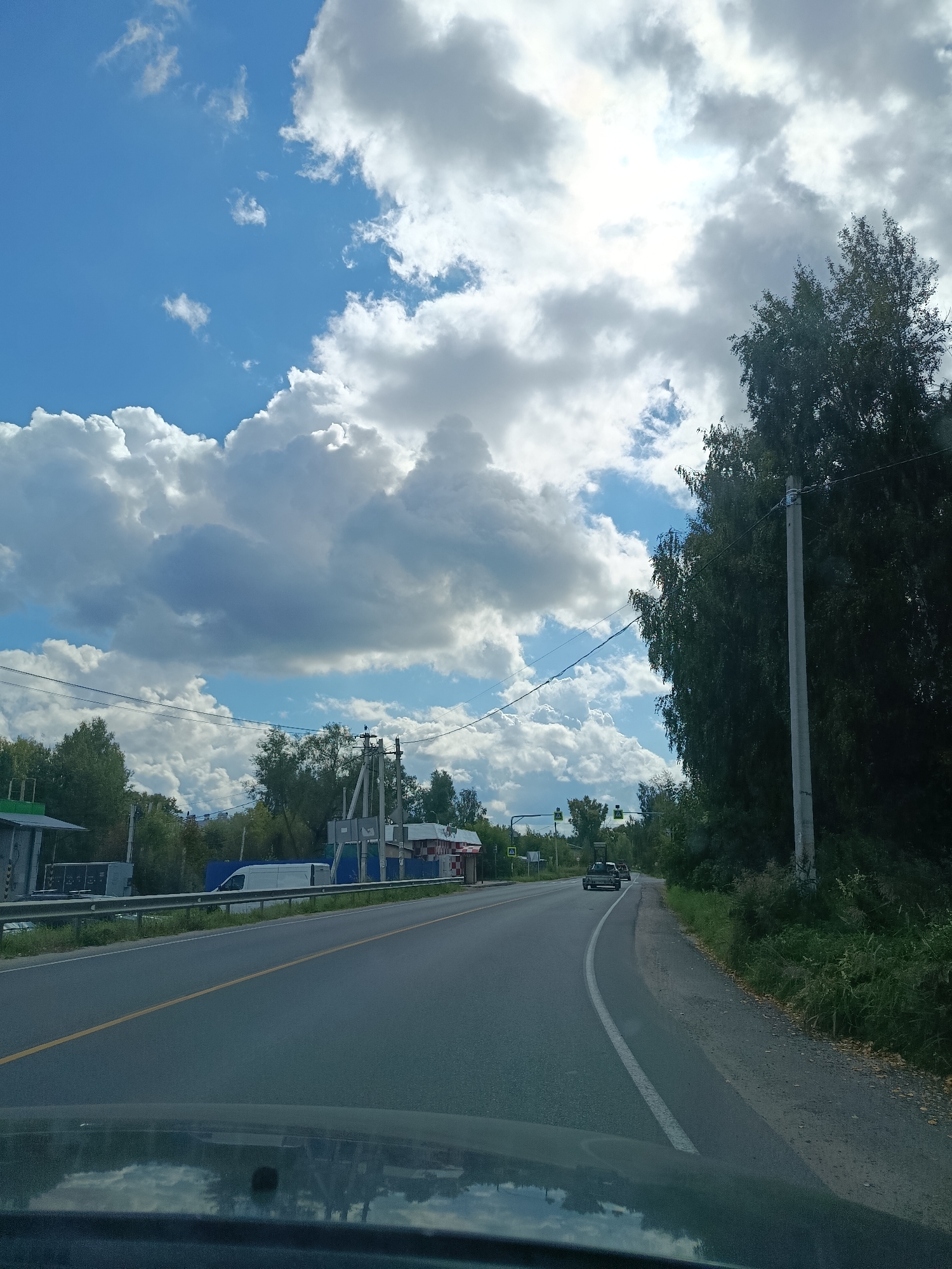 The image captures a road scene in the vicinity of Снопок Новый, Russia, under a bright, partly cloudy sky. The perspective is from within a vehicle, looking forward and slightly to the right. A paved road with a yellow dividing line stretches into the distance, curving gently to the right. On the left side of the road, there's a white van parked next to a building with a green awning. Further down the road, a cluster of utility poles and some trees are visible. The right side of the road is bordered by a dense line of trees and vegetation, with a concrete utility pole prominently placed in the foreground. The sky is filled with large, fluffy white and gray clouds, with patches of blue sky peeking through, and the sun is partially obscured by the clouds, creating a bright, diffused light. A dark-colored car is visible in the distance on the road, moving away from the viewer. The reflection of the sky and clouds is visible on the lower part of the image, likely from the vehicle's dashboard or windshield.