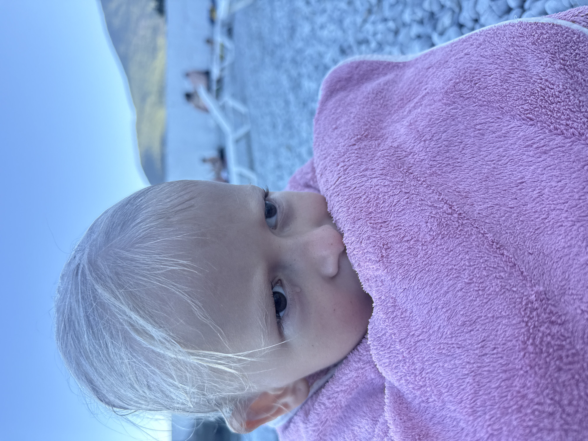 A close-up shot focuses on a young child wrapped in a pink towel, their face turned to the side with a tear visible on their cheek. The child has light-colored hair and appears to be looking away from the camera. In the background, out of focus, are lounge chairs and people on a beach, with a body of water and mountains in the distance under a clear blue sky. The setting suggests a beach or coastal area.