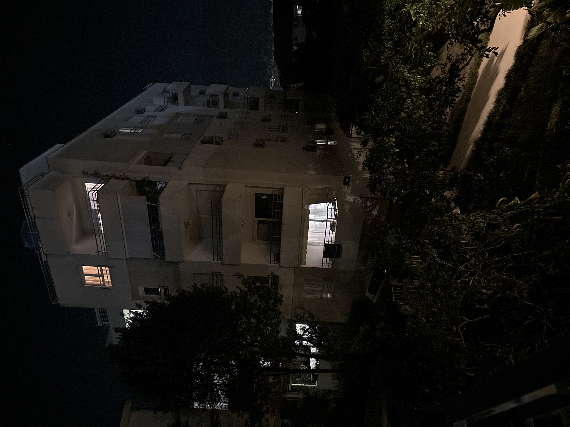 An exterior, low-angle, nighttime, medium shot captures the side of a multi-story apartment building illuminated by artificial light. The building is constructed of light-colored concrete and features multiple balconies with dark railings. Several windows are visible, with some emitting a warm, interior glow, suggesting occupancy. A large, dark tree with dense foliage stands to the left of the building, partially obscuring it. Below the building, a walkway or path is visible, casting long shadows from the surrounding vegetation. The ground slopes downwards from the building towards the path, covered in dark, leafy bushes. The night sky is black, indicating the absence of significant ambient light from the moon or stars. In the distance, some faint lights suggest the presence of other structures or a populated area, consistent with the city of Budva, Montenegro. No people are visible in the frame.